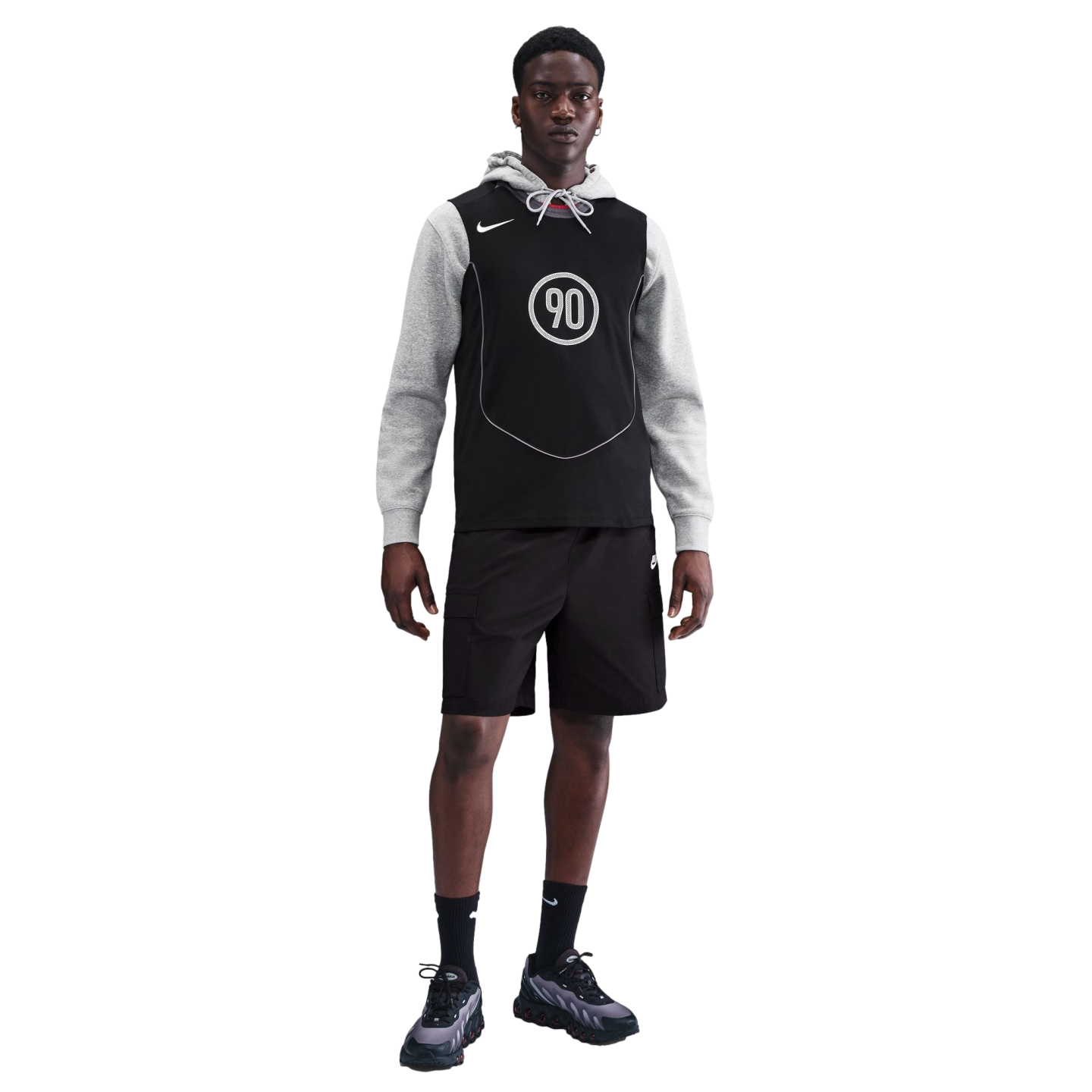 A Black man in a gray hoodie, black Nike jersey with "90" logo, black cargo shorts, and sneakers, standing against a solid black background.