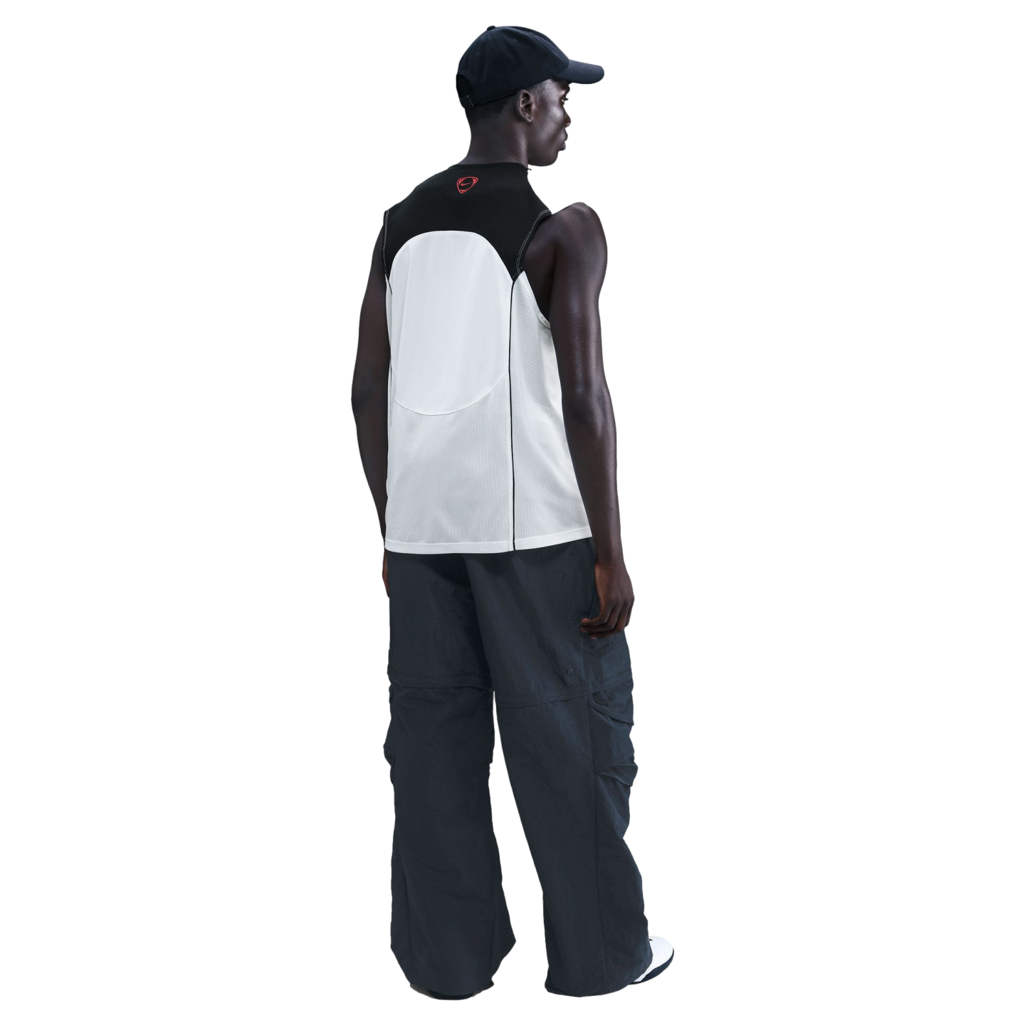 Back view of a person wearing a black cap, a white and black sleeveless mesh top, and dark grey baggy pants, standing against a black background.