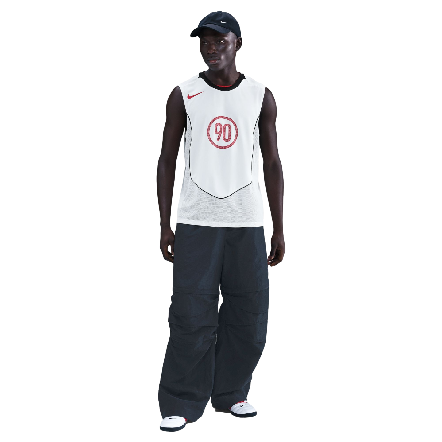 A man wearing a white Nike "90" jersey, extremely baggy dark pants, a navy cap, and white sneakers against a black background.