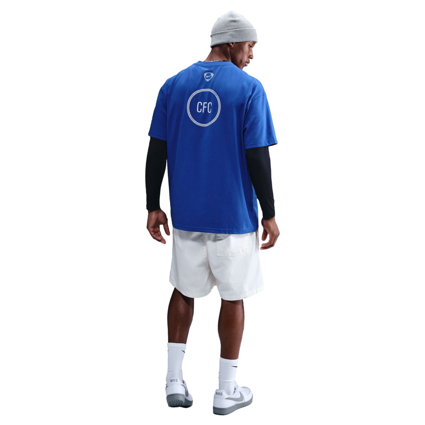 Rear view of a man wearing a blue CFC t-shirt over black long sleeves, white shorts, a grey beanie, and Nike sneakers against a black background.