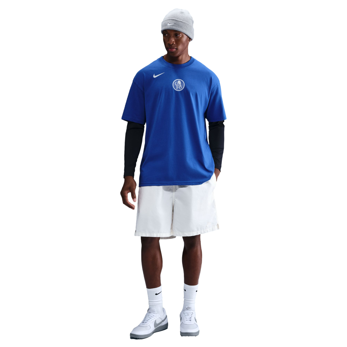 A person in a blue Nike Chelsea FC t-shirt over black long sleeves, white shorts, and a grey beanie stands against a black background.
