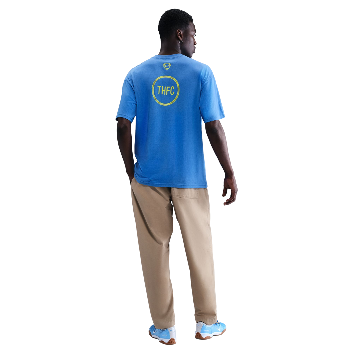 Back view of a man wearing a blue Nike t-shirt with a yellow "THFC" graphic, tan pants, and blue sneakers against a black background.