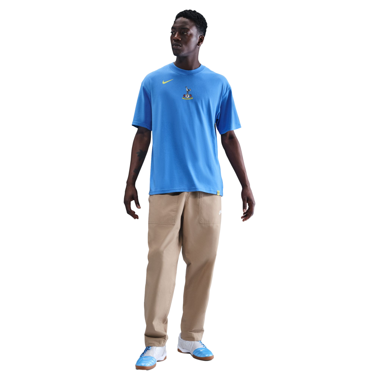 Full-length portrait of a man wearing a light blue Nike Tottenham Hotspur t-shirt, tan trousers, and blue and white sneakers, standing against a solid black background.
