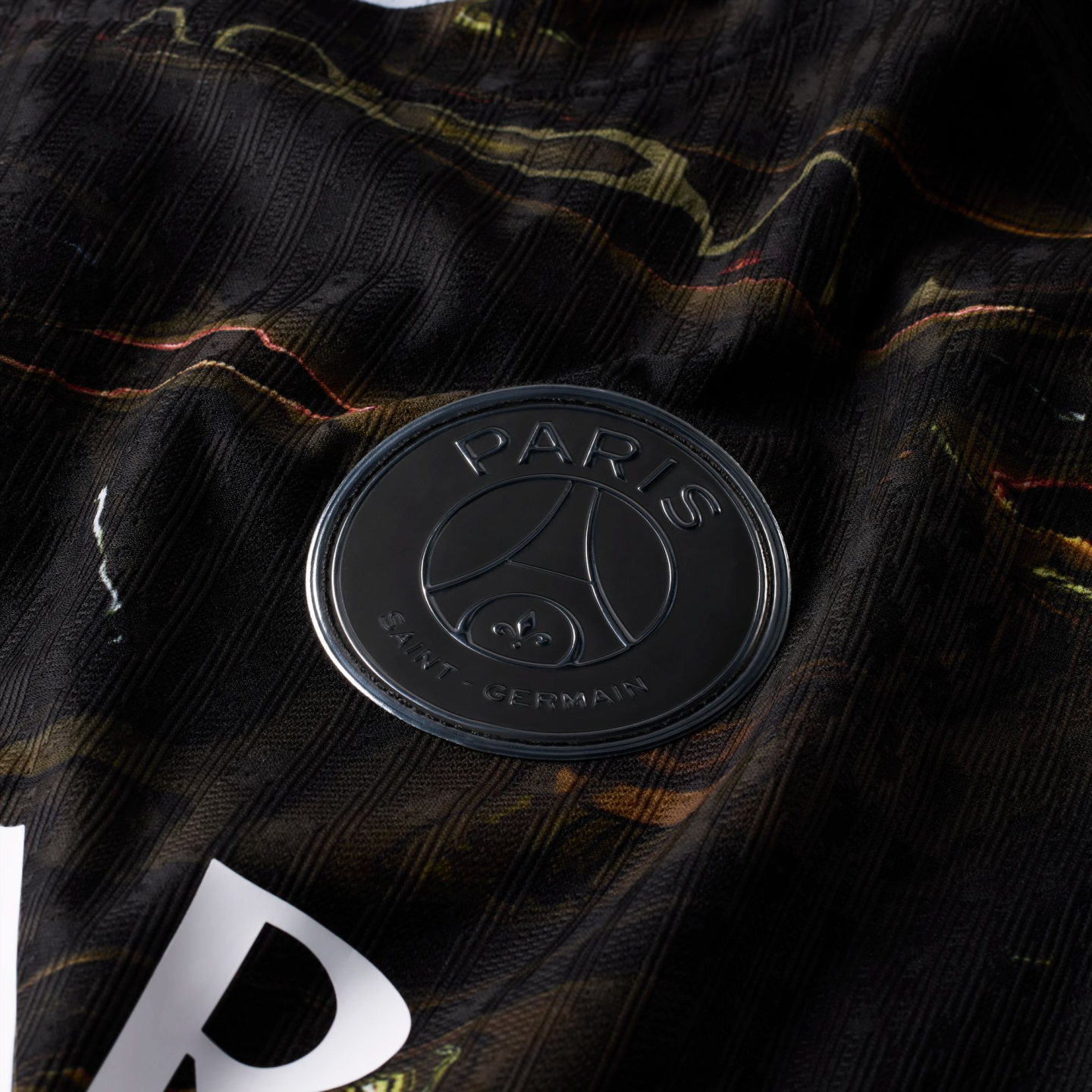 A close-up photograph of a matte black Paris Saint-Germain (PSG) football club badge. The circular badge, featuring raised lettering and the team's iconic Eiffel Tower and fleur-de-lis logo, is set against a dark, ribbed fabric jersey with marbled gold an