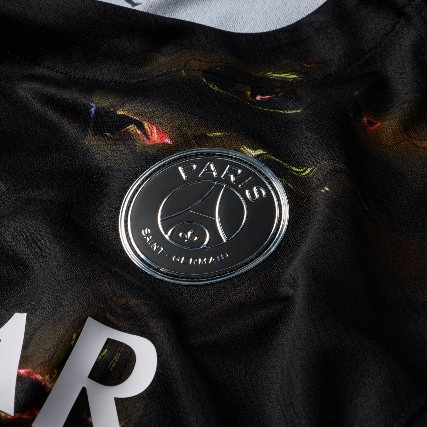 Close-up of a black sports jersey featuring a metallic silver Paris Saint-Germain club crest. The fabric has an abstract, multicolored pattern with hints of red and green.