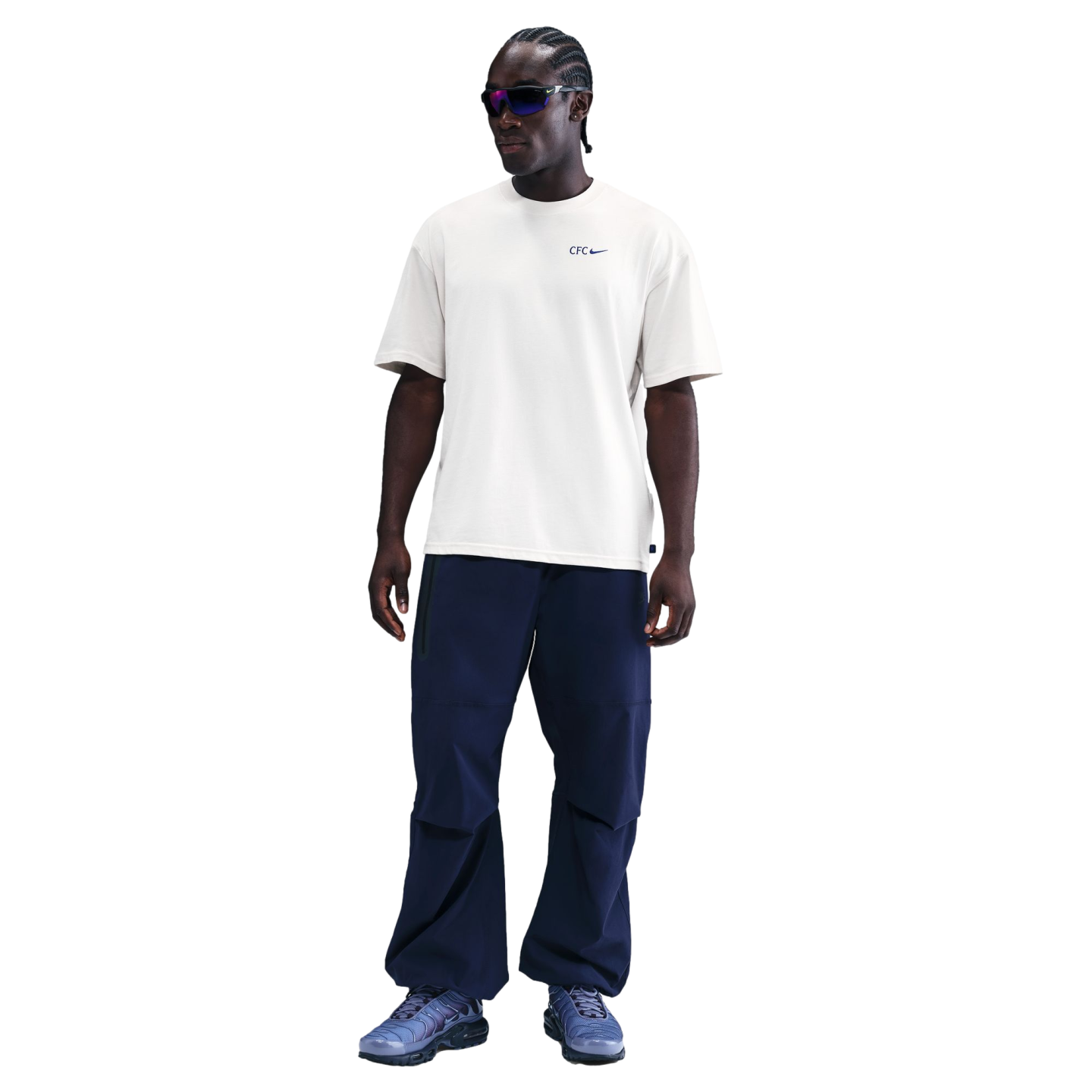 A man wearing a cream Nike CFC t-shirt, navy cargo pants, and blue sneakers against a black background.