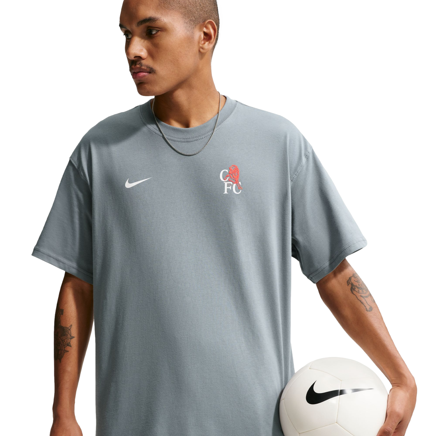 A person with arm tattoos wearing a grey Nike Chelsea FC t-shirt holds a soccer ball against a black background.