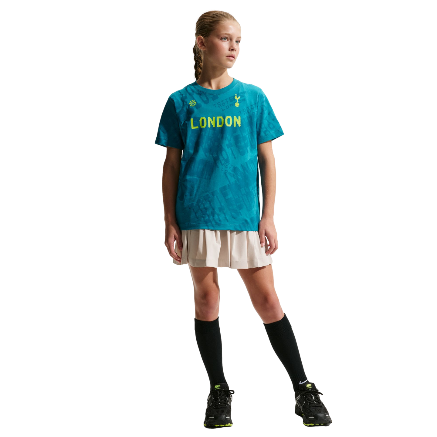 A young girl in a teal "LONDON" t-shirt, beige pleated skirt, and black sneakers poses against a black background.