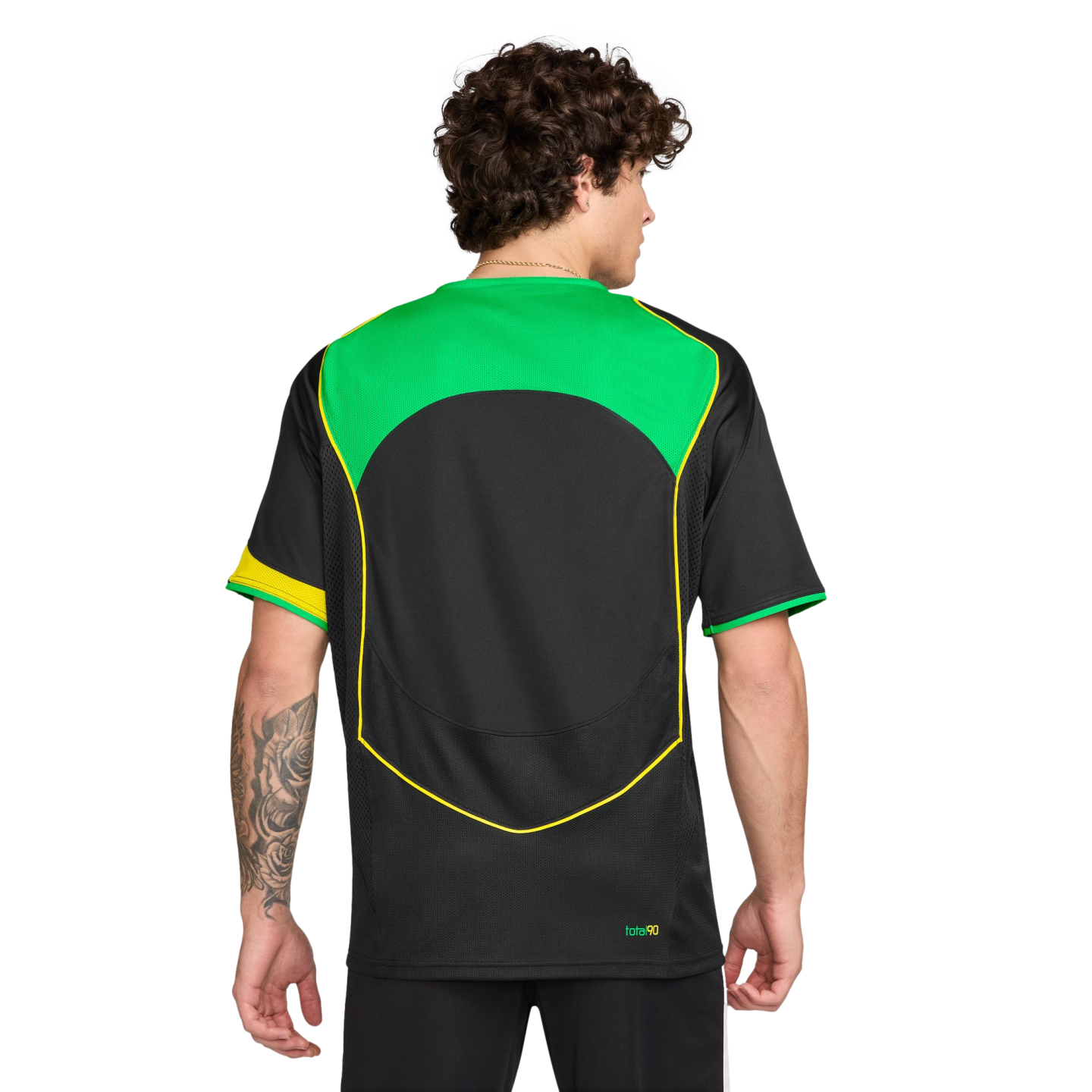 Rear view of a man with curly hair wearing a black soccer jersey with green and yellow accents. He has a tattoo on his left forearm. Plain white background.