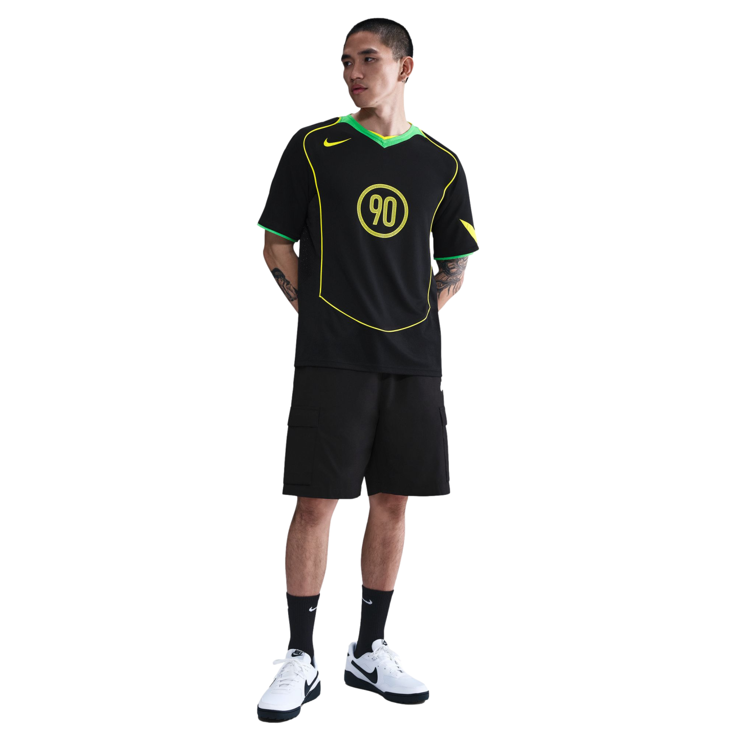 A man poses in a black Nike T90 soccer jersey, black cargo shorts, and white sneakers against a white background.
