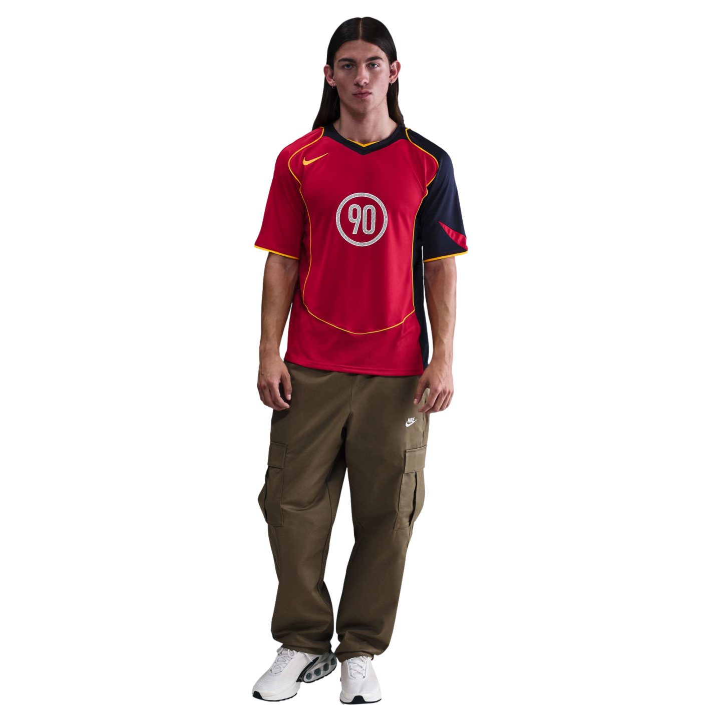 Man with long dark hair wearing a red Nike Total 90 jersey, olive green cargo pants, and white sneakers.