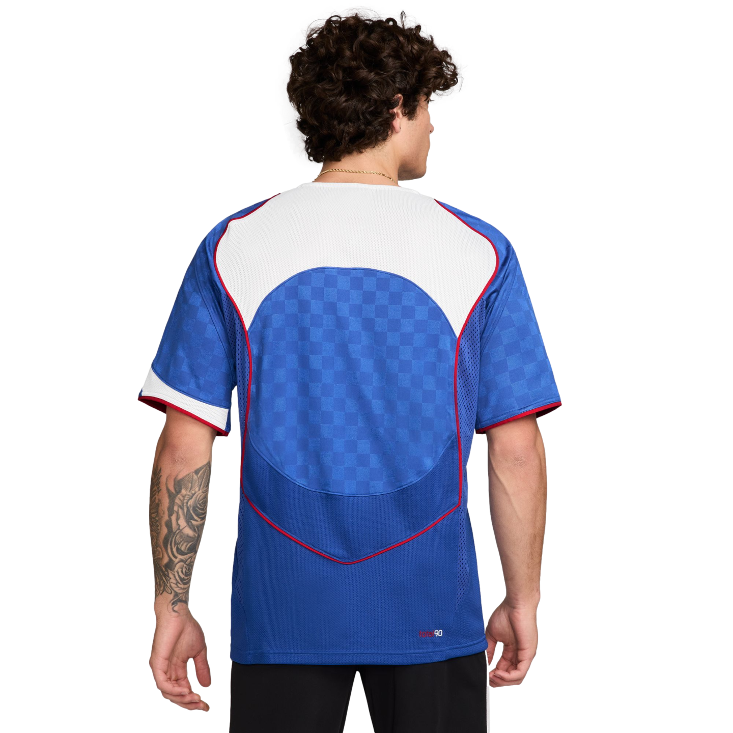 Back view of a man with curly hair wearing a blue soccer jersey with a white yoke, red trim, and a checkered pattern on the back.