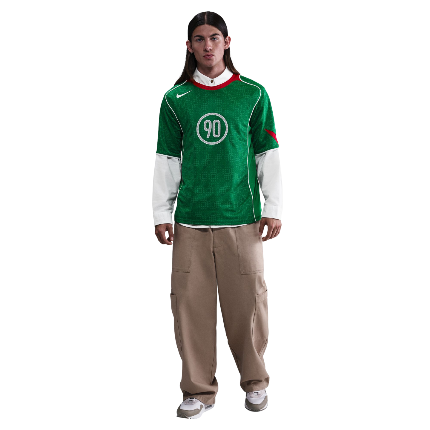 A person with long dark hair wears a green Nike soccer jersey over a white button-down shirt, baggy tan cargo pants, and sneakers, standing against a plain white background.