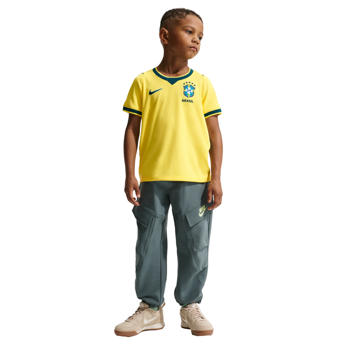 A full-body studio portrait of a young boy standing against a black background. He is wearing a yellow Nike Brazil soccer jersey with green trim, grey cargo-style jogger pants, and tan sneakers.