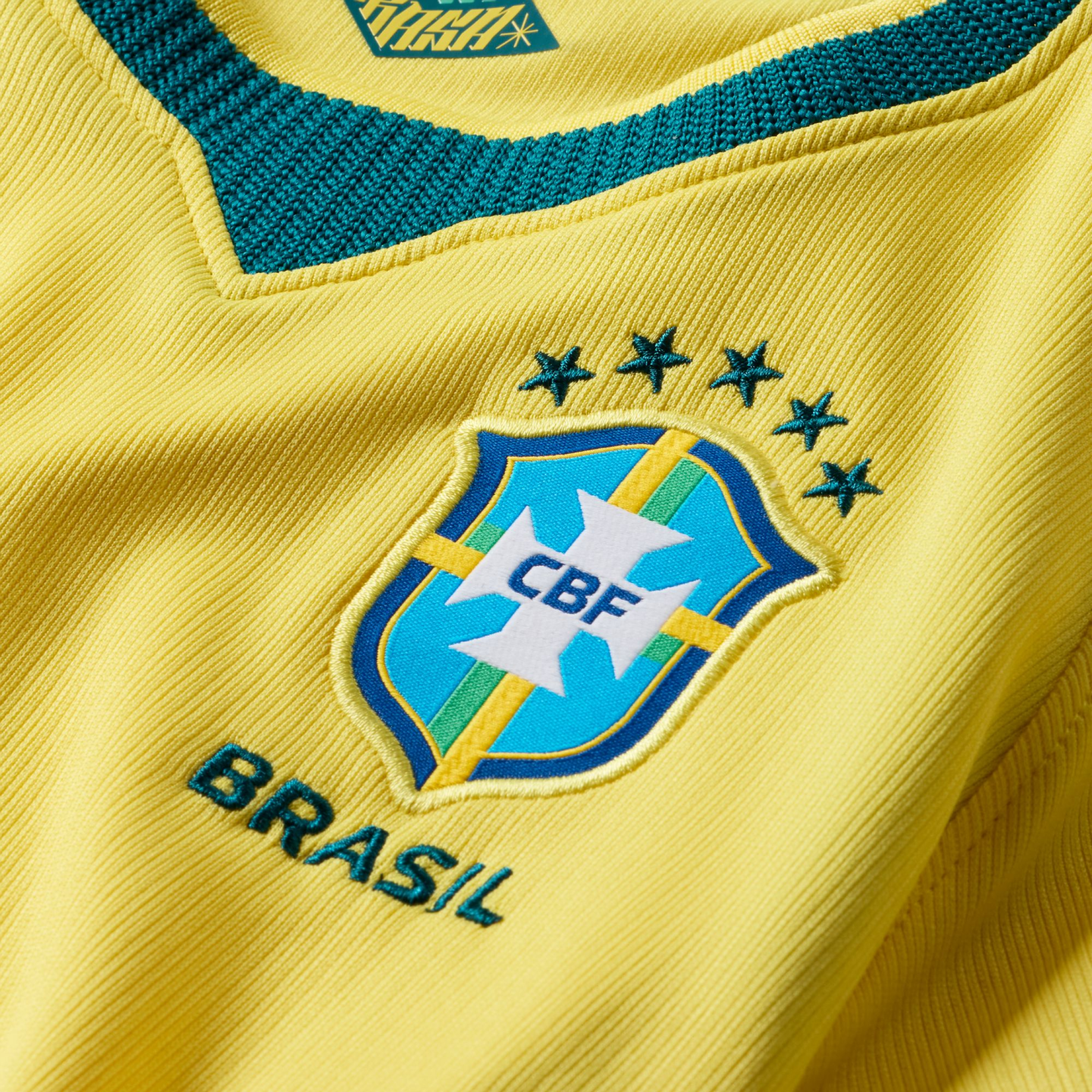 A close-up of the yellow ribbed fabric of a Brazilian national football jersey. The blue and white CBF crest is featured in the center, topped by five green stars and with the word "BRASIL" embroidered in green below it. A dark green, textured V-neck coll