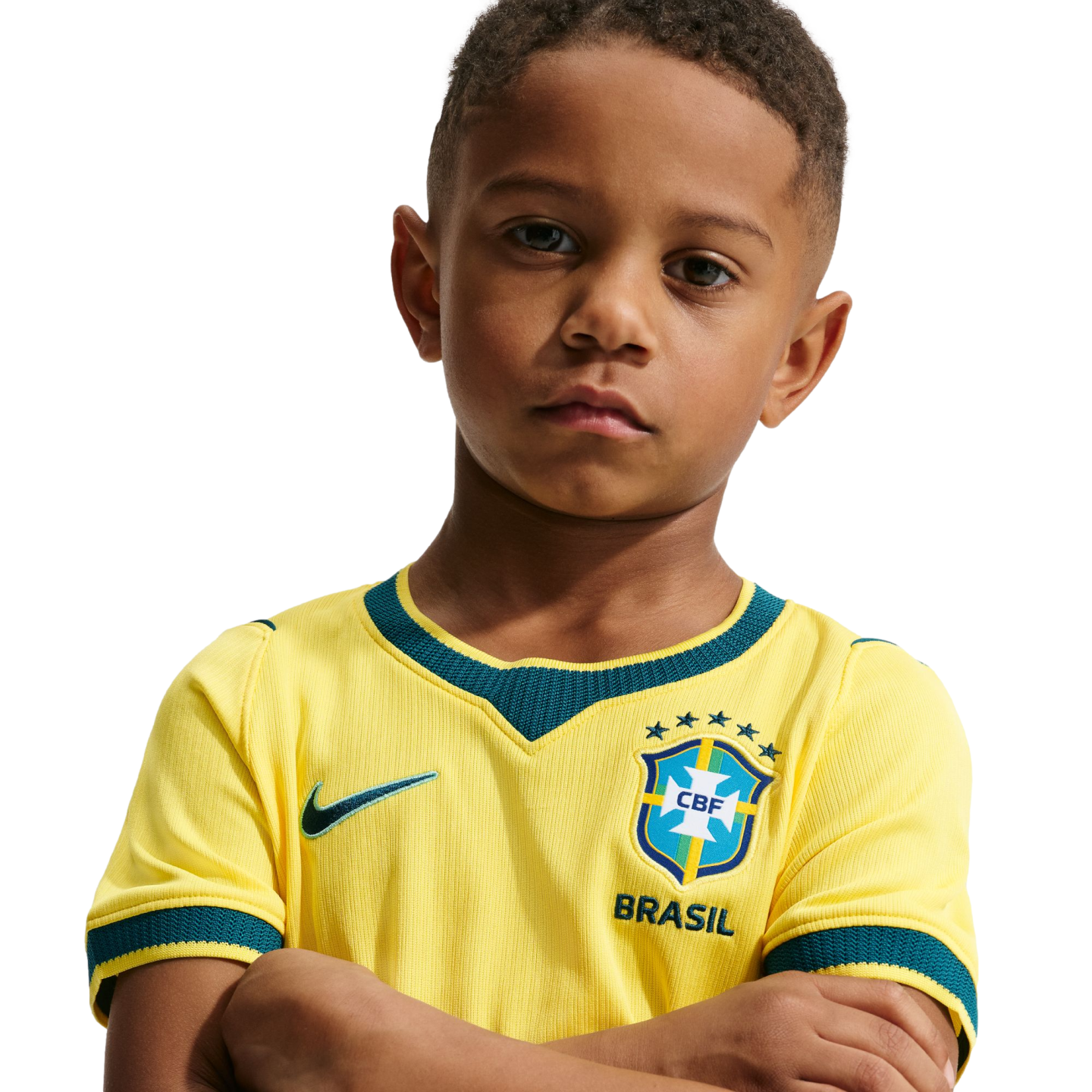 A young boy with a serious expression and arms crossed, wearing a yellow Brazil national soccer team jersey against a solid black background.