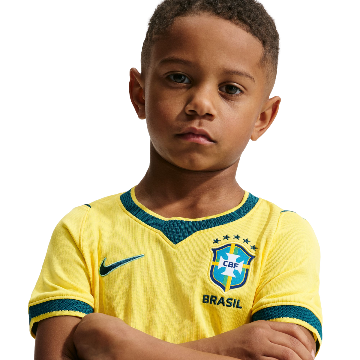 A young boy with a serious expression and arms crossed, wearing a yellow Brazil national soccer team jersey against a solid black background.