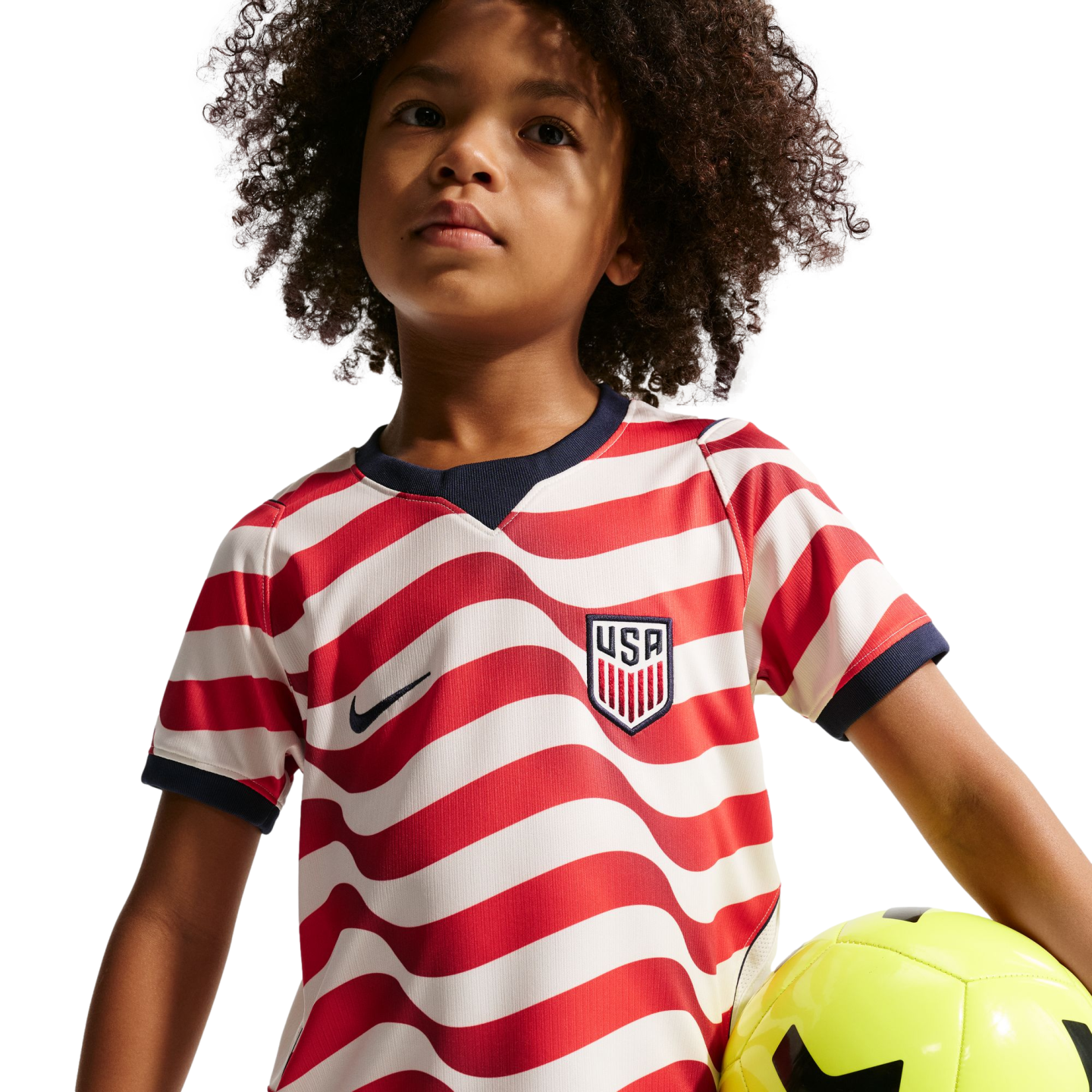 A young child with curly hair wearing a red and white striped USA soccer jersey holding a yellow soccer ball against a black background.