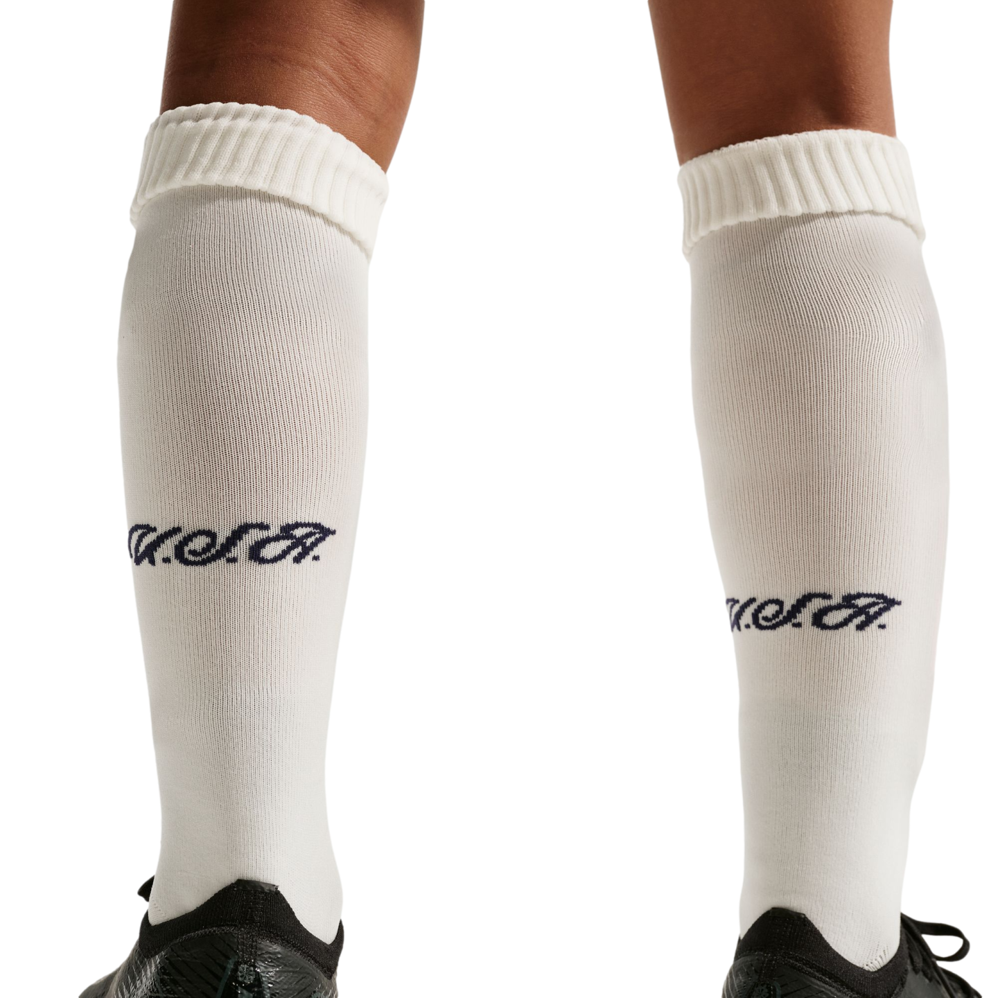 A close-up of a person's legs wearing white knee-high socks with a navy blue "USA" script logo on the calves and black athletic shoes, set against a solid black background.