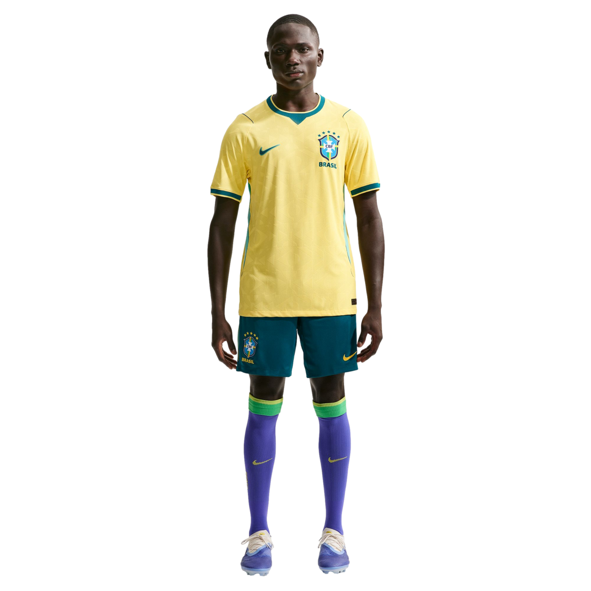 A full-length studio portrait of a man modeling the Brazil national football team uniform against a solid black background. He is wearing the team's iconic yellow jersey with green accents, blue shorts, purple socks with green bands at the top, and light