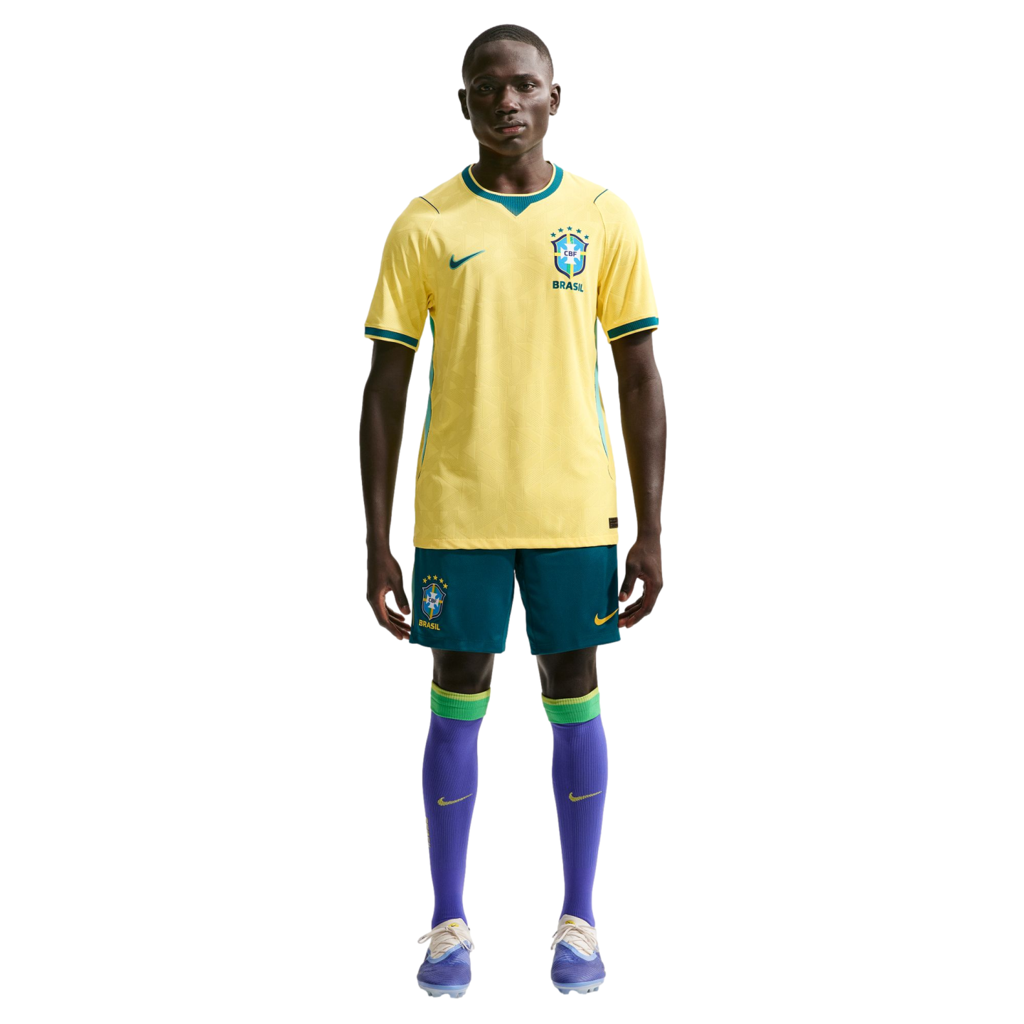 A full-length studio portrait of a man modeling the Brazil national football team uniform against a solid black background. He is wearing the team's iconic yellow jersey with green accents, blue shorts, purple socks with green bands at the top, and light