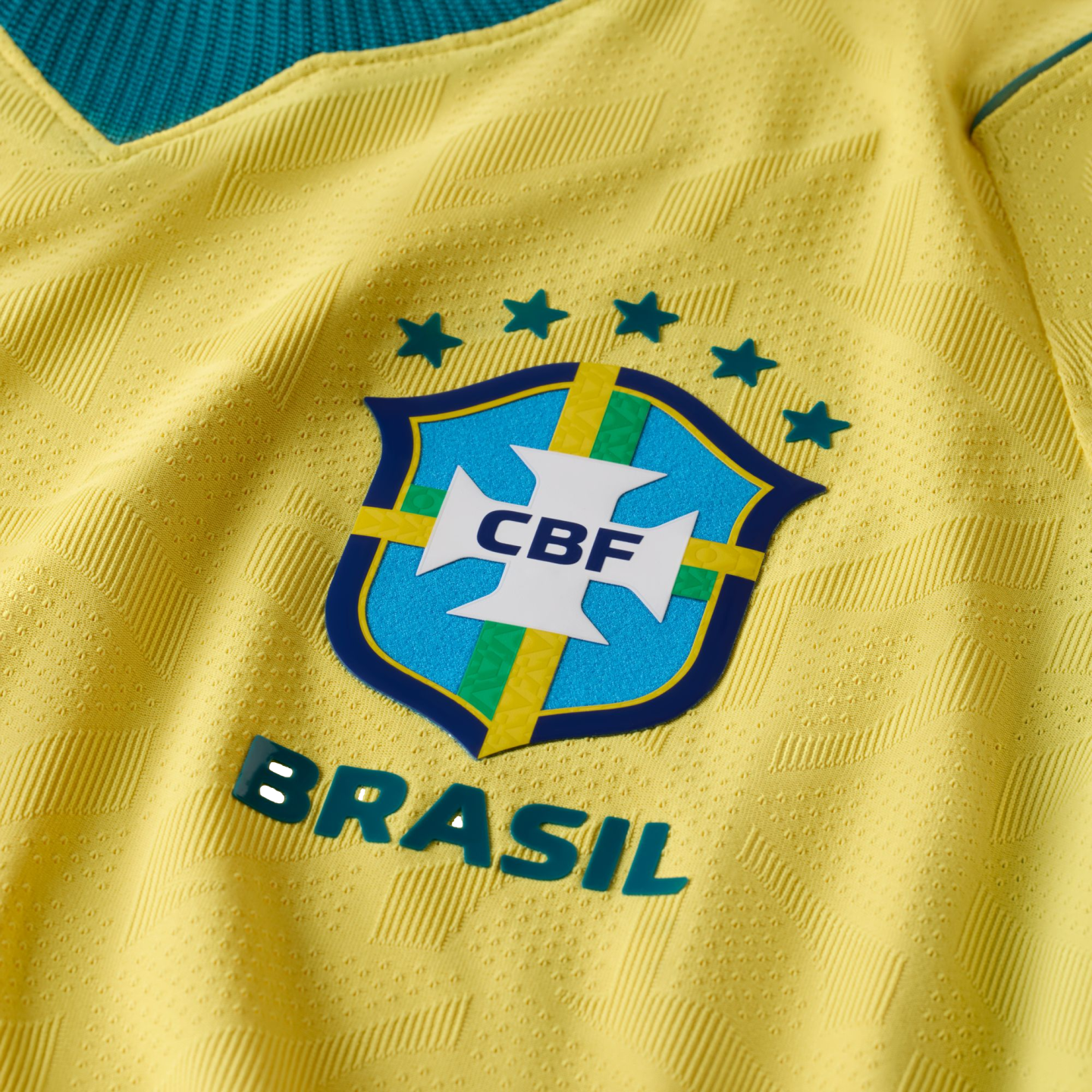 A close-up of a yellow Brazilian national football team jersey, featuring the blue CBF crest, five teal stars above it, and the word "BRASIL" in raised letters below. The yellow fabric has an intricate, textured geometric pattern.