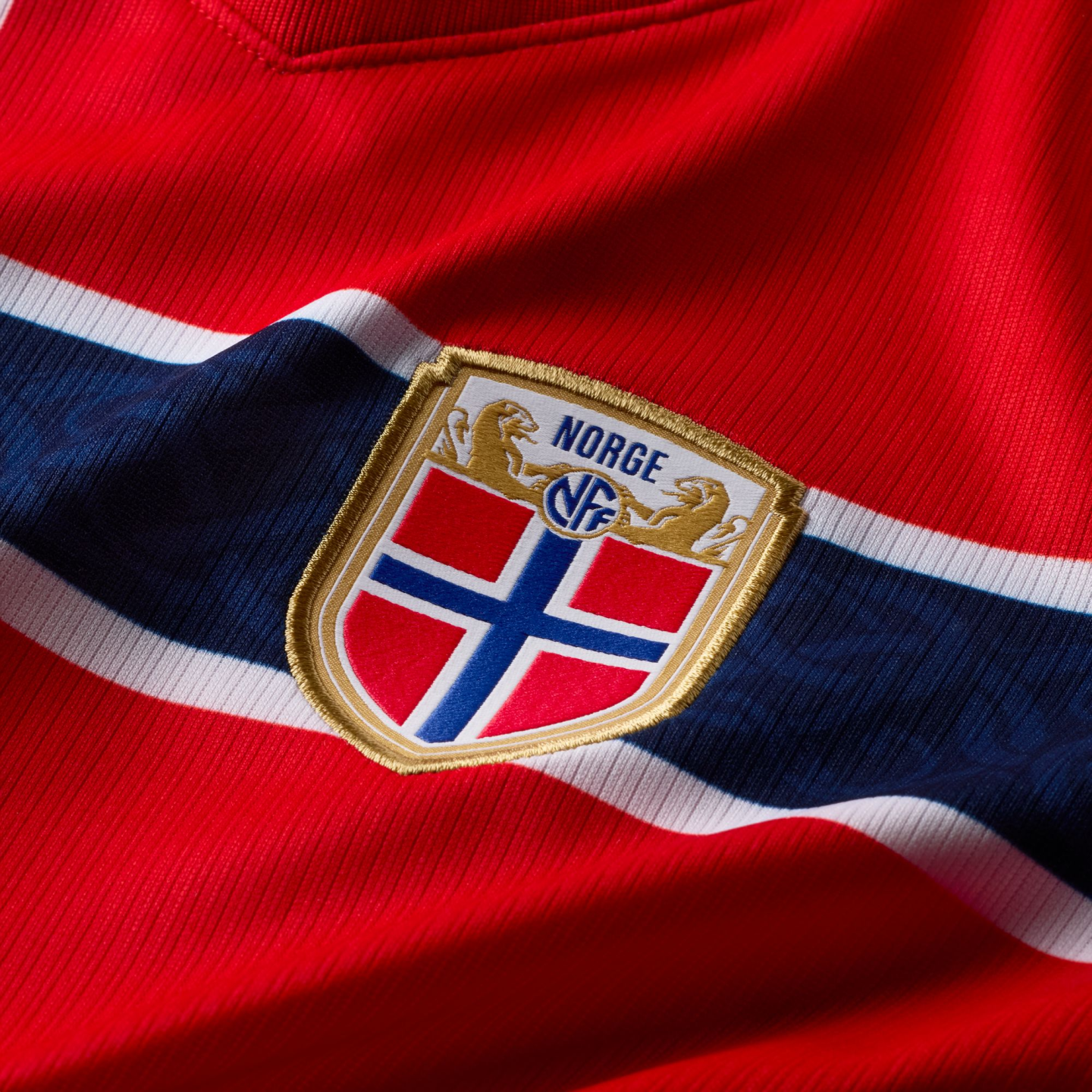 A close-up of a red Norway national team football jersey. A horizontal navy blue stripe with white borders runs across the fabric. Centered on the stripe is the Norwegian Football Federation (NFF) crest, featuring a gold-bordered shield with the Norwegian