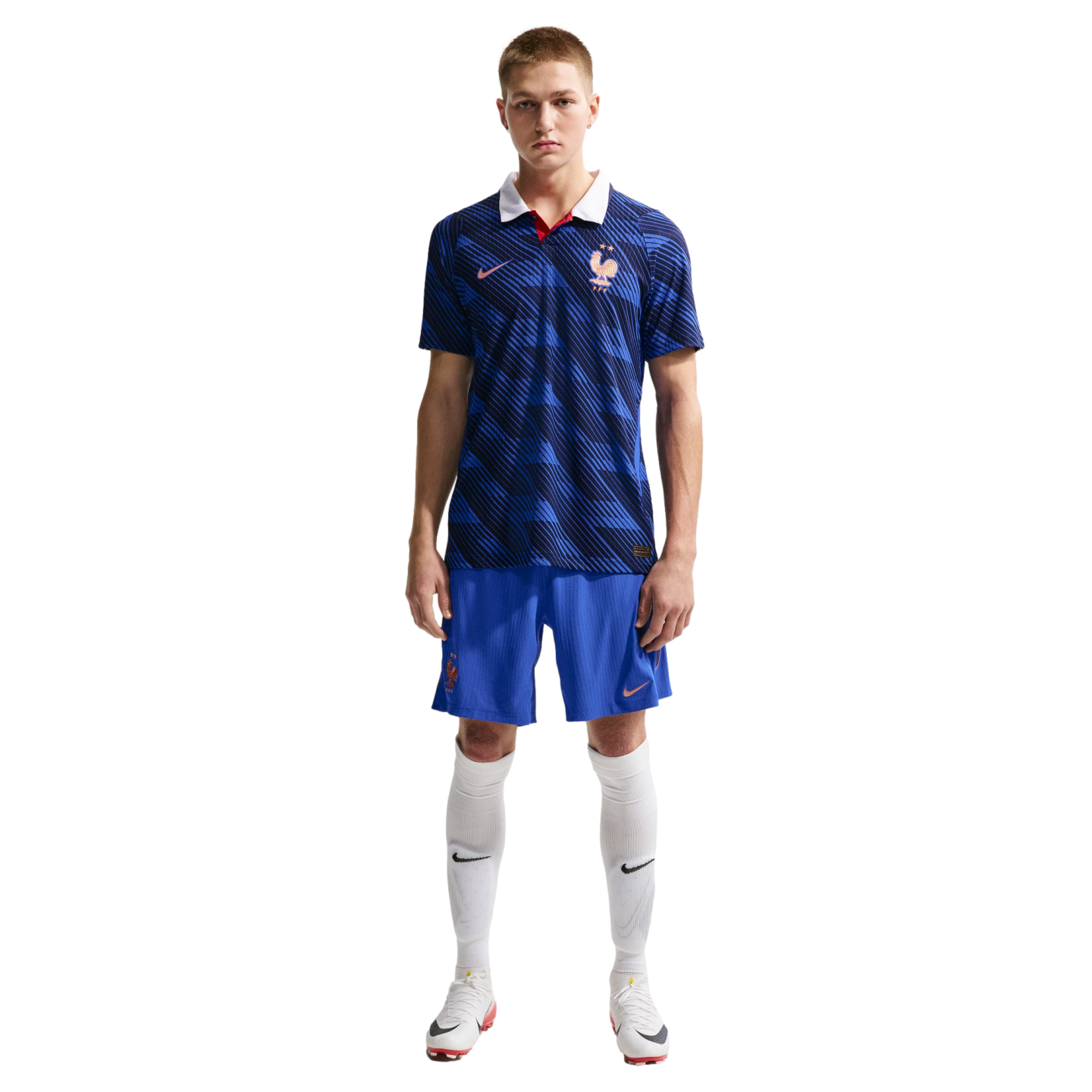 A man in a blue patterned France national football kit with a white collar, blue shorts, and white socks, standing against a black background.