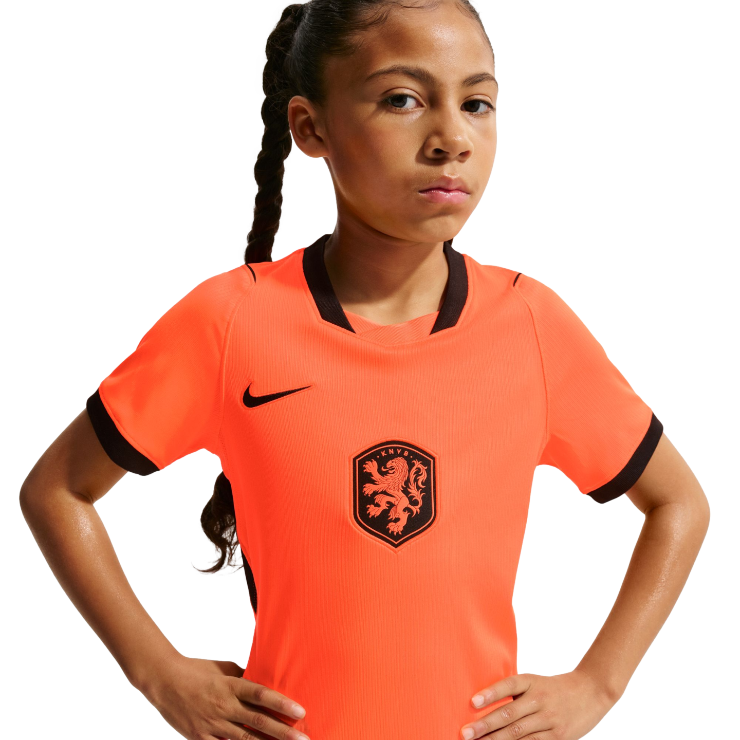 A young girl with a long braid and a serious expression poses with hands on her hips, wearing an orange Nike Netherlands national team soccer jersey against a solid black background.