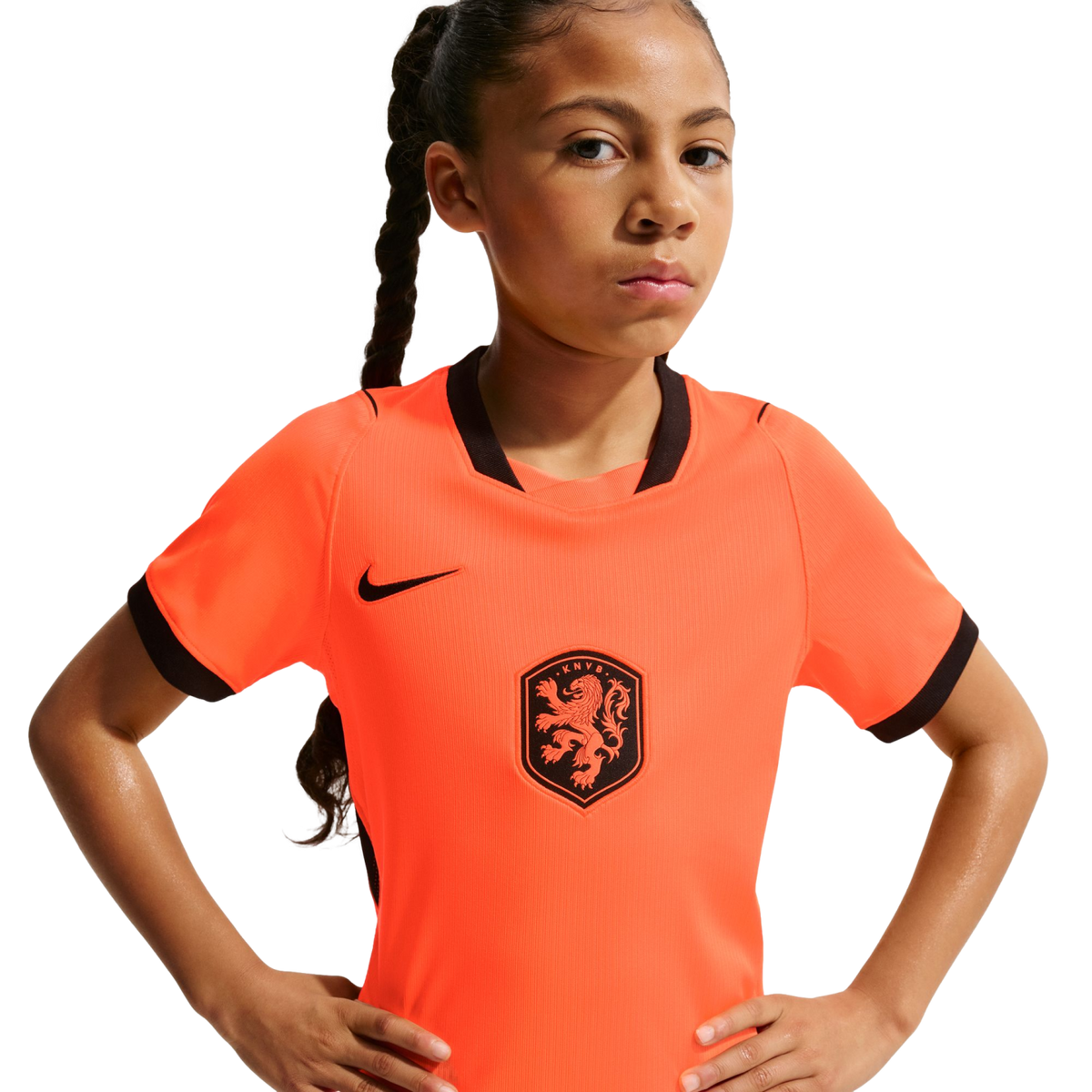 A young girl with a long braid and a serious expression poses with hands on her hips, wearing an orange Nike Netherlands national team soccer jersey against a solid black background.