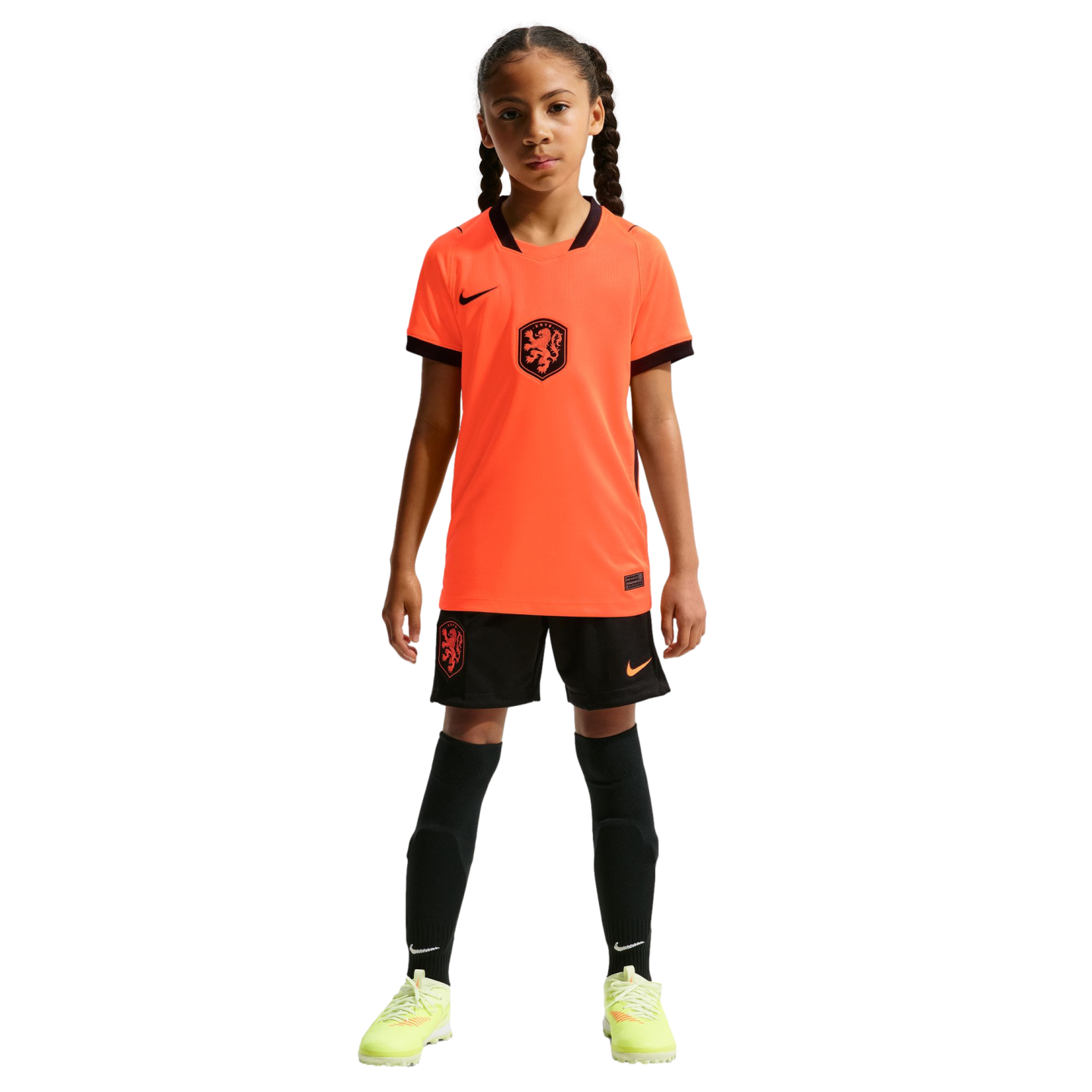 A young girl stands facing forward in a full Netherlands national soccer kit, including a bright orange Nike jersey, black shorts, tall black socks, and neon yellow cleats, against a solid black background.