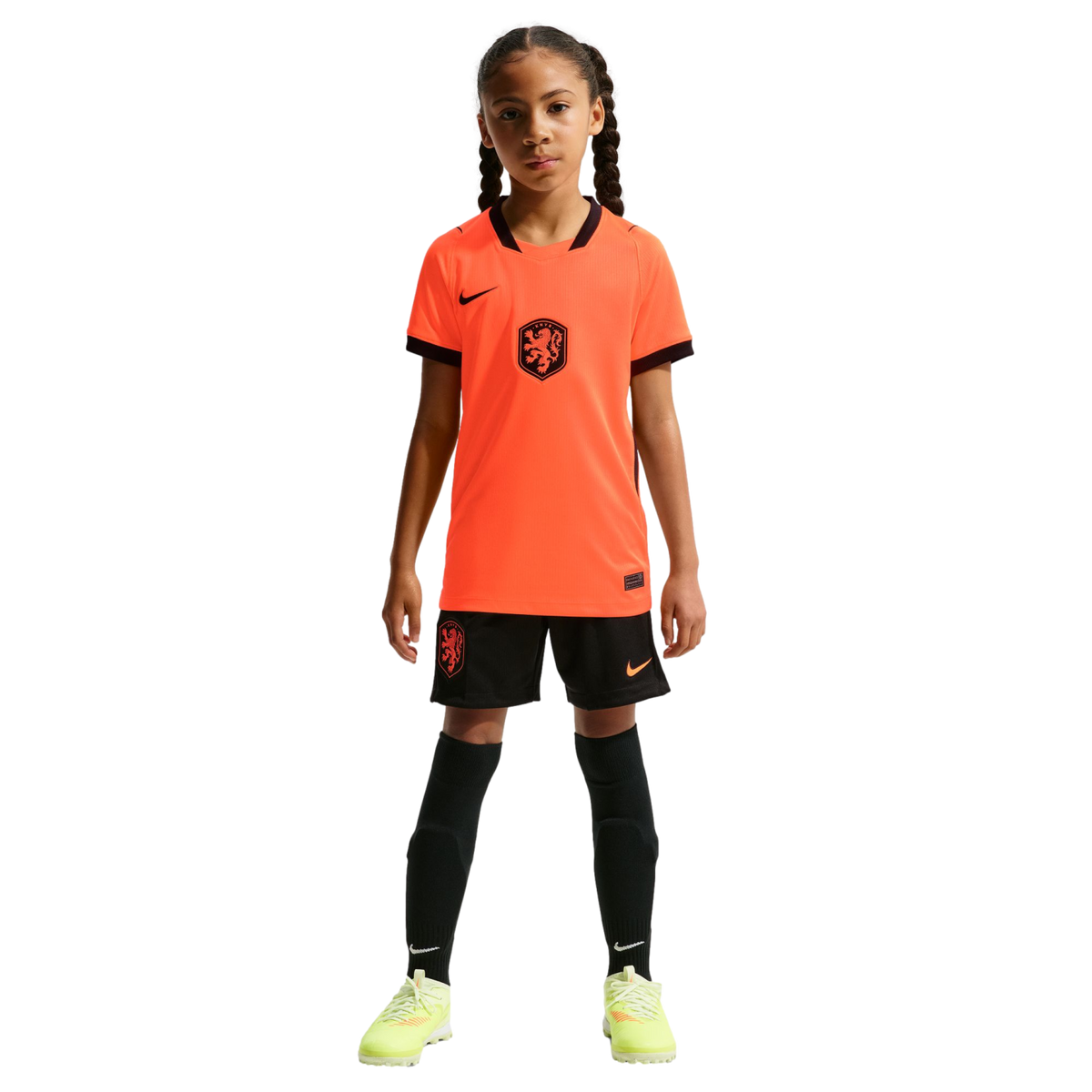 A young girl stands facing forward in a full Netherlands national soccer kit, including a bright orange Nike jersey, black shorts, tall black socks, and neon yellow cleats, against a solid black background.