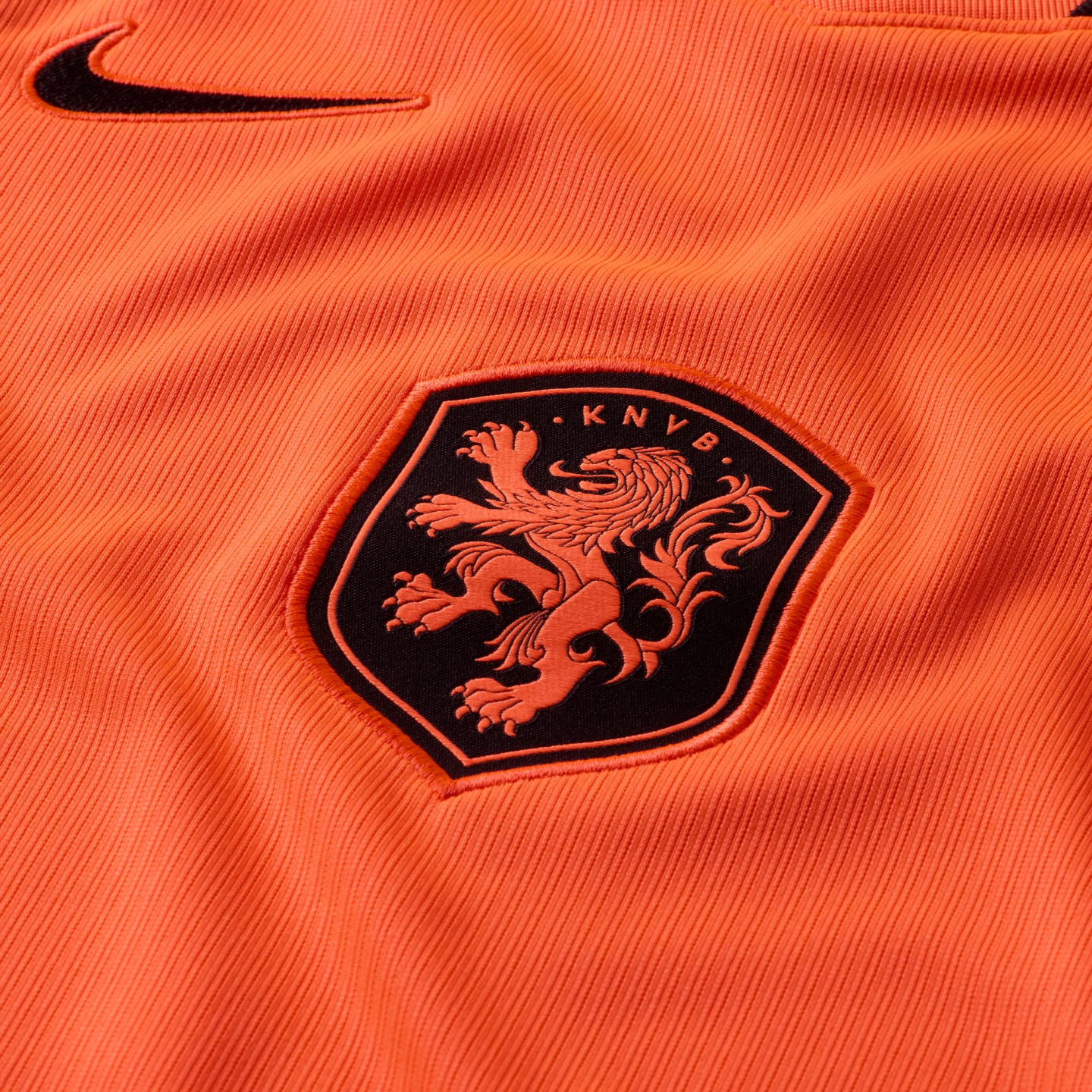 A close-up of an orange ribbed sports jersey featuring a black Nike swoosh in the upper left corner and the Netherlands national football team crest, a shield with an orange lion on a black background and the letters "KNVB."