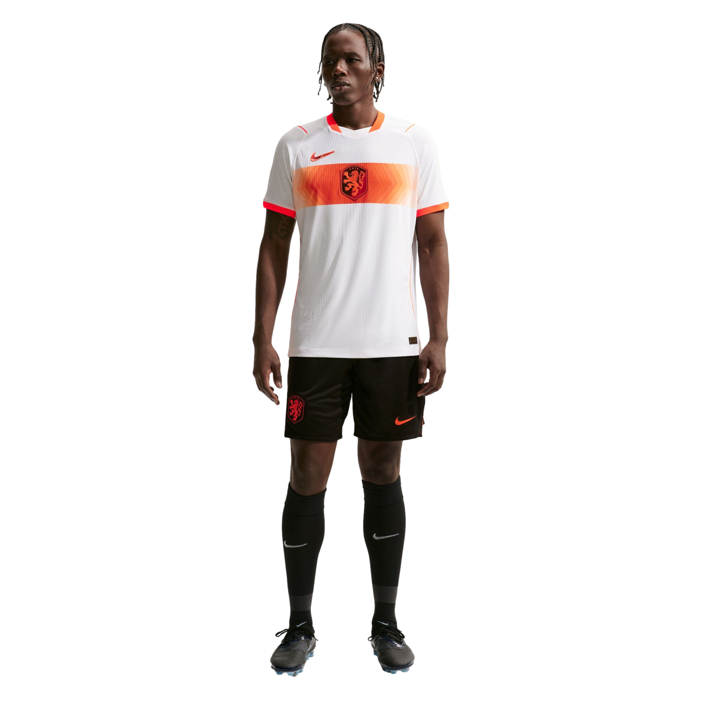 A full-length portrait of a male athlete wearing a Netherlands national soccer team away kit, standing against a solid black background. He is wearing a white jersey with an orange horizontal stripe across the chest, black shorts, and black knee-high sock