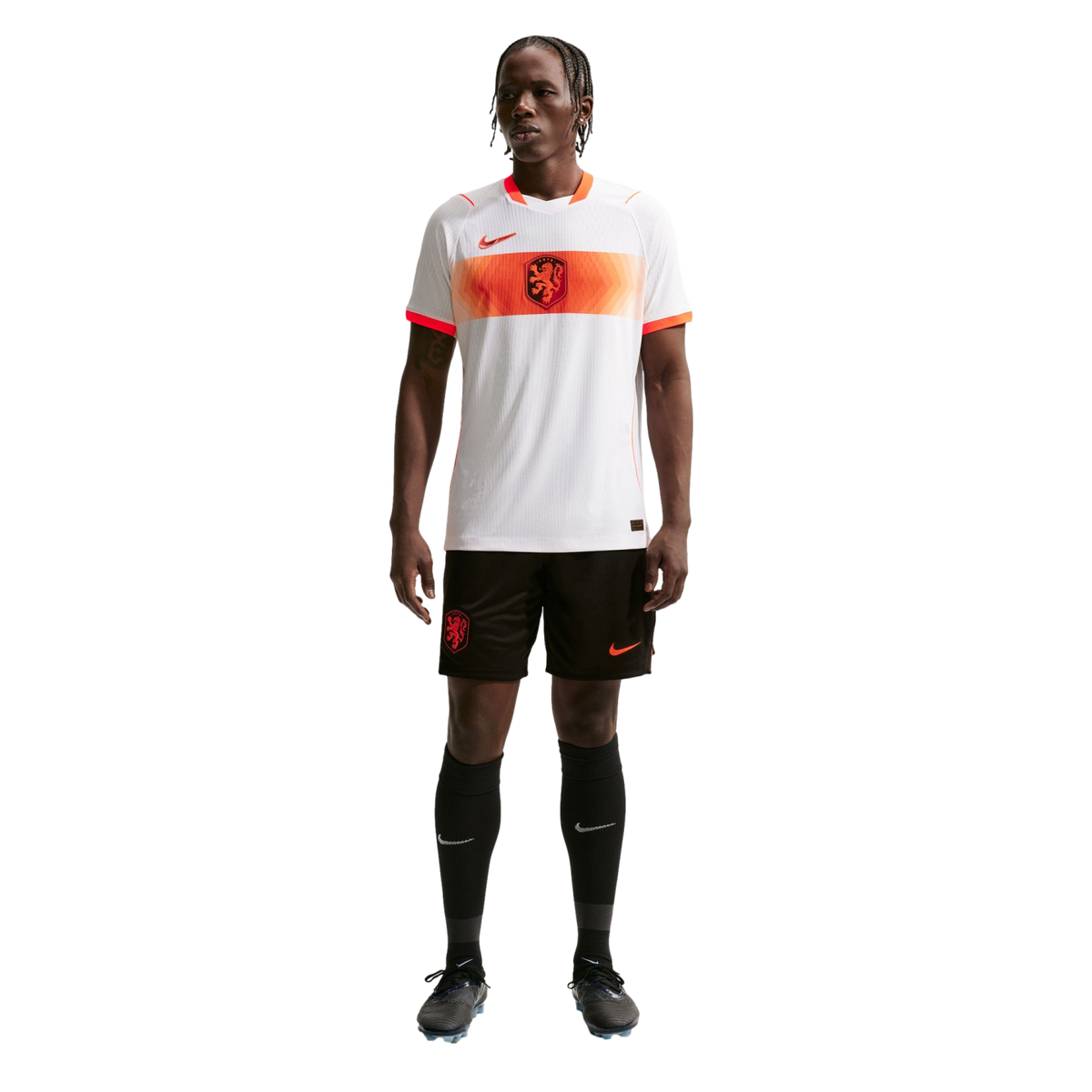 A full-length portrait of a male athlete wearing a Netherlands national soccer team away kit, standing against a solid black background. He is wearing a white jersey with an orange horizontal stripe across the chest, black shorts, and black knee-high sock