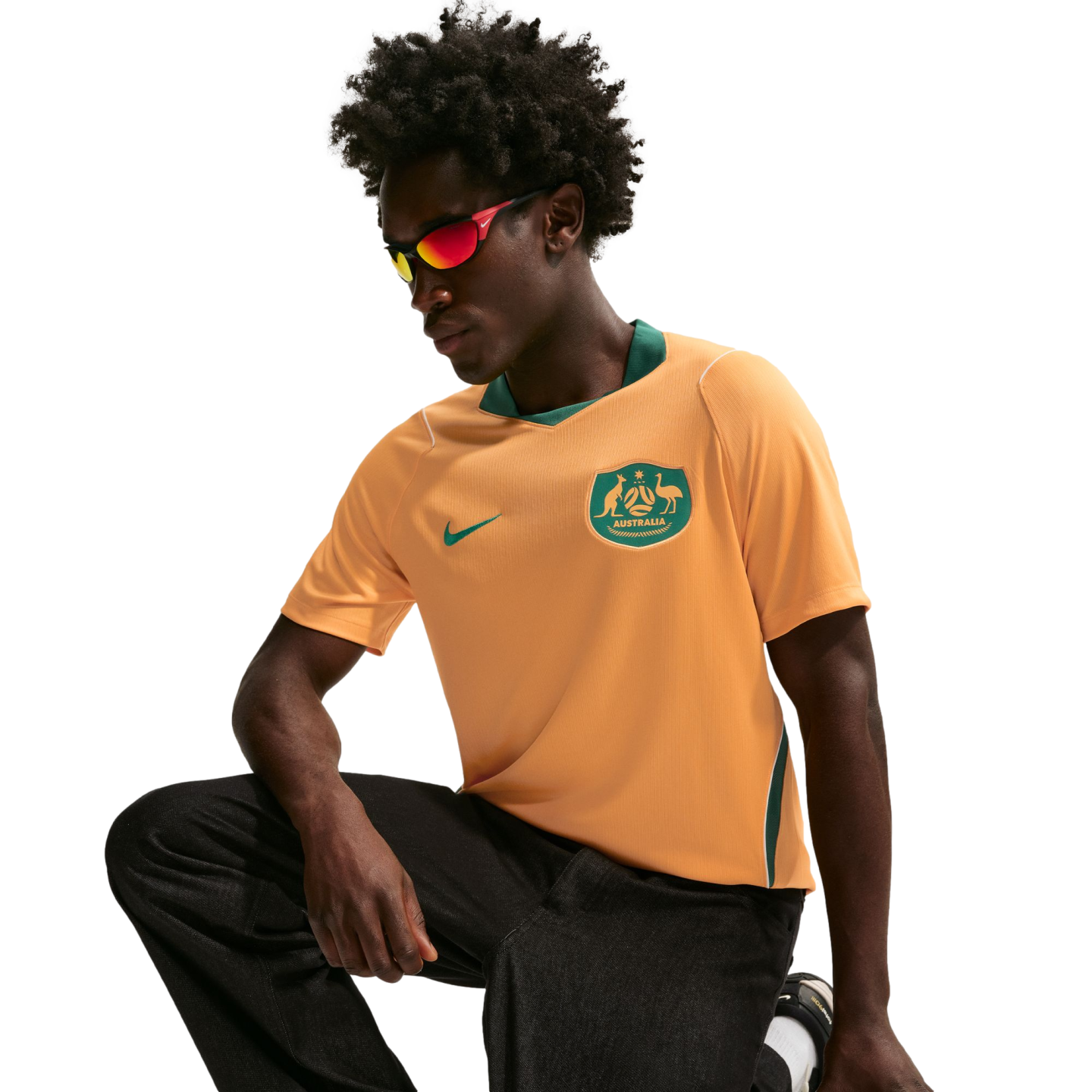 A man wearing an orange and green Australia national soccer jersey and red-tinted sunglasses crouches against a black background.