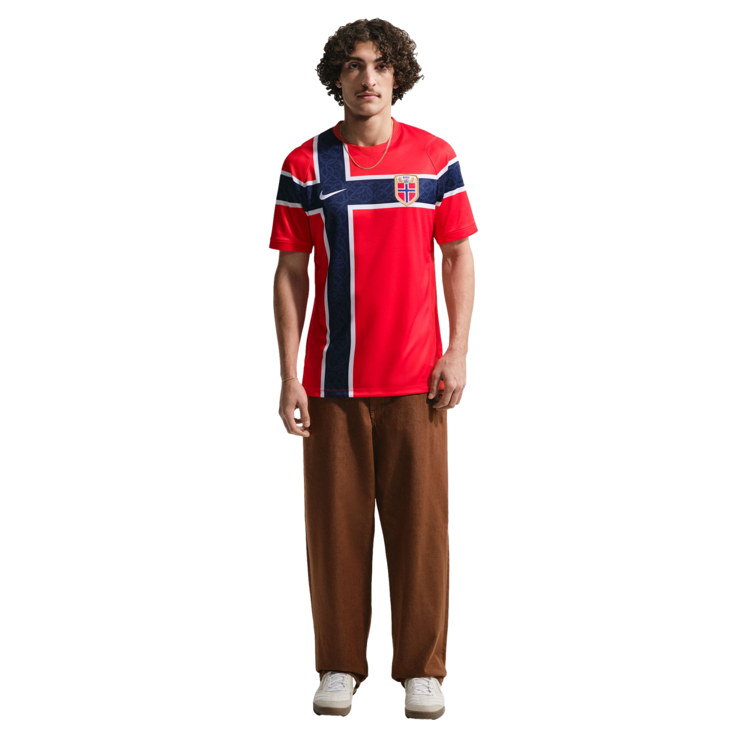 A young man stands against a black background wearing a red Nike Norway national team soccer jersey, brown baggy trousers, and white sneakers.