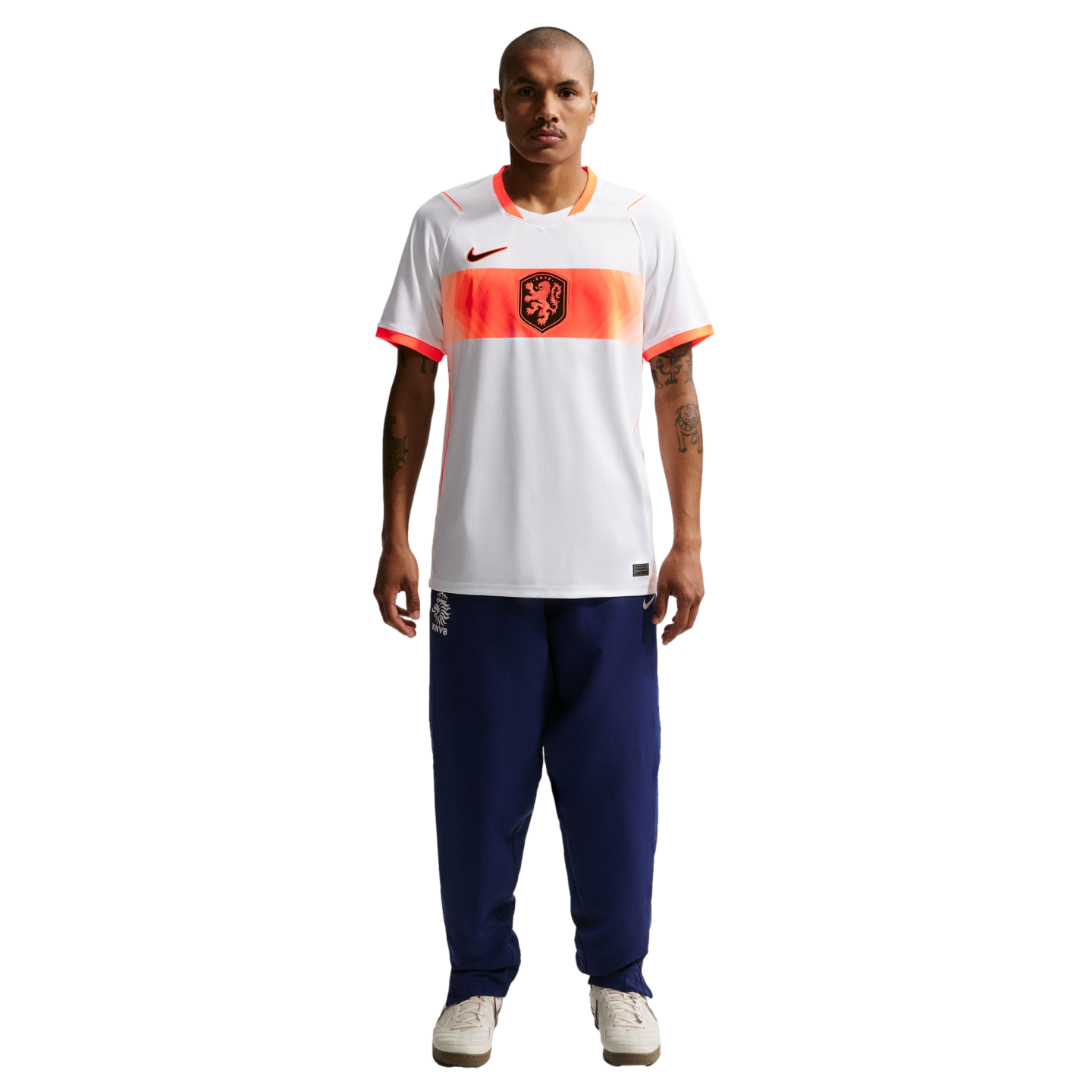 A full-length studio portrait of a man standing against a black background. He is wearing a white Nike soccer jersey with an orange horizontal band across the chest that features a black lion crest. He is also wearing navy blue tracksuit pants and off-whi