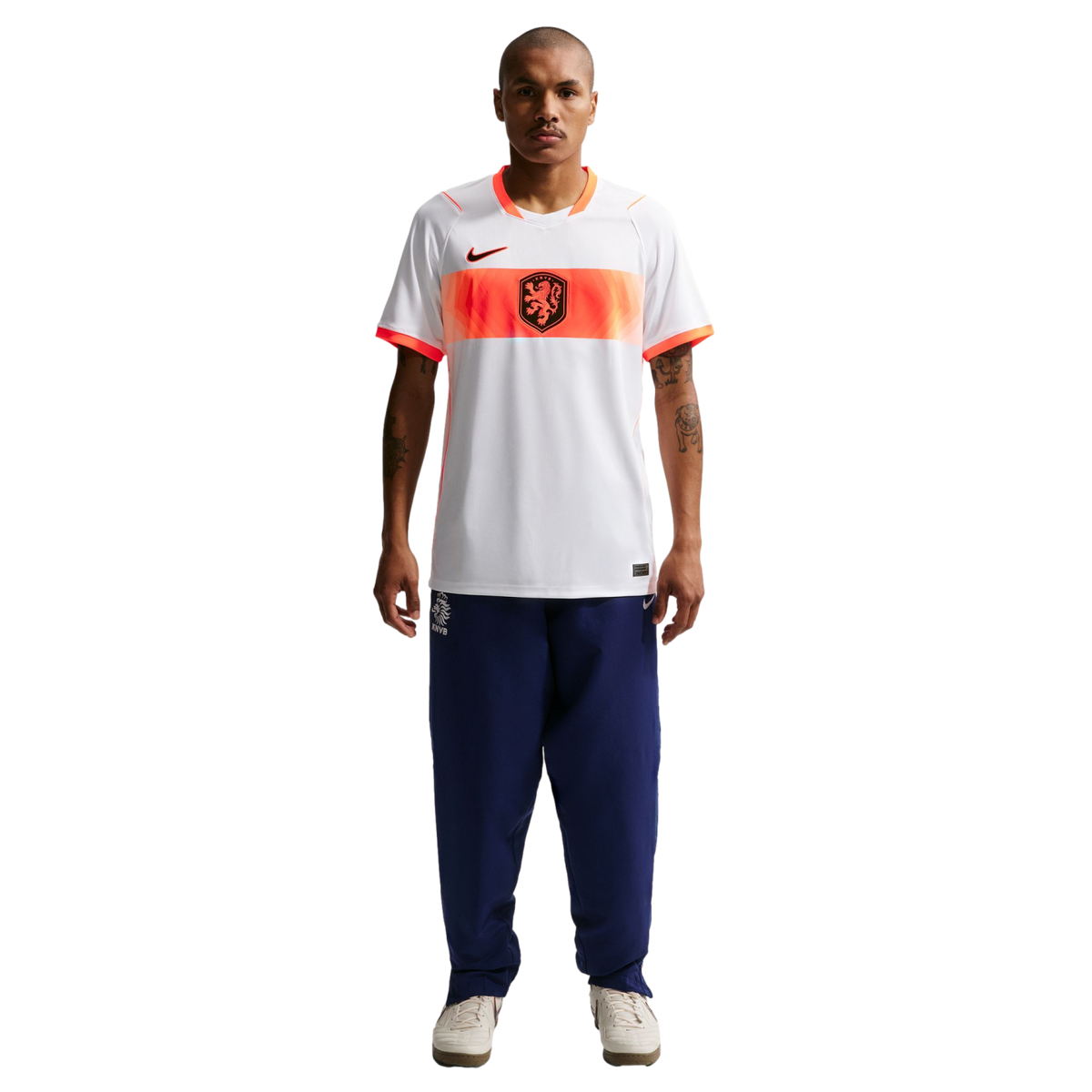 A full-length studio portrait of a man standing against a black background. He is wearing a white Nike soccer jersey with an orange horizontal band across the chest that features a black lion crest. He is also wearing navy blue tracksuit pants and off-whi