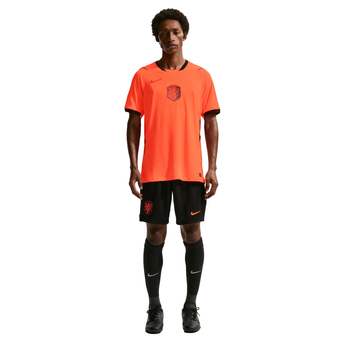 A male model stands against a black background wearing a bright orange Netherlands national team soccer jersey with black trim, matching black shorts, high black socks, and black cleats.
