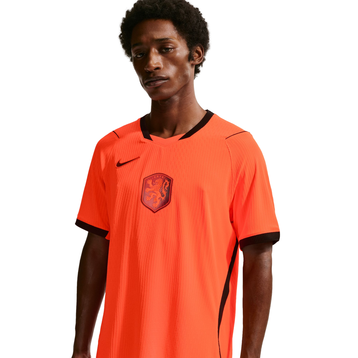 A young man with dark skin and curly hair wears a bright orange Netherlands national team home jersey. The jersey has a black V-neck collar, sleeve trim, and a black Nike swoosh on the chest, with the KNVB lion crest in the center. He is positioned agains
