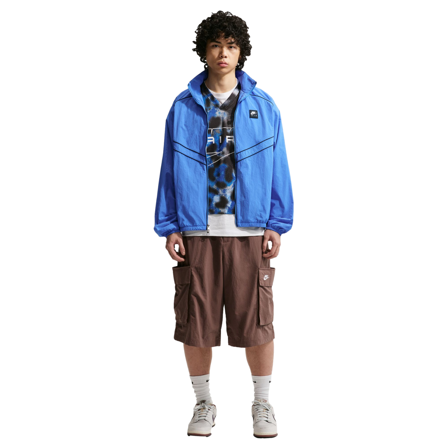 A man with curly hair poses in a blue windbreaker, a patterned top, brown cargo shorts, and sneakers against a plain white background.