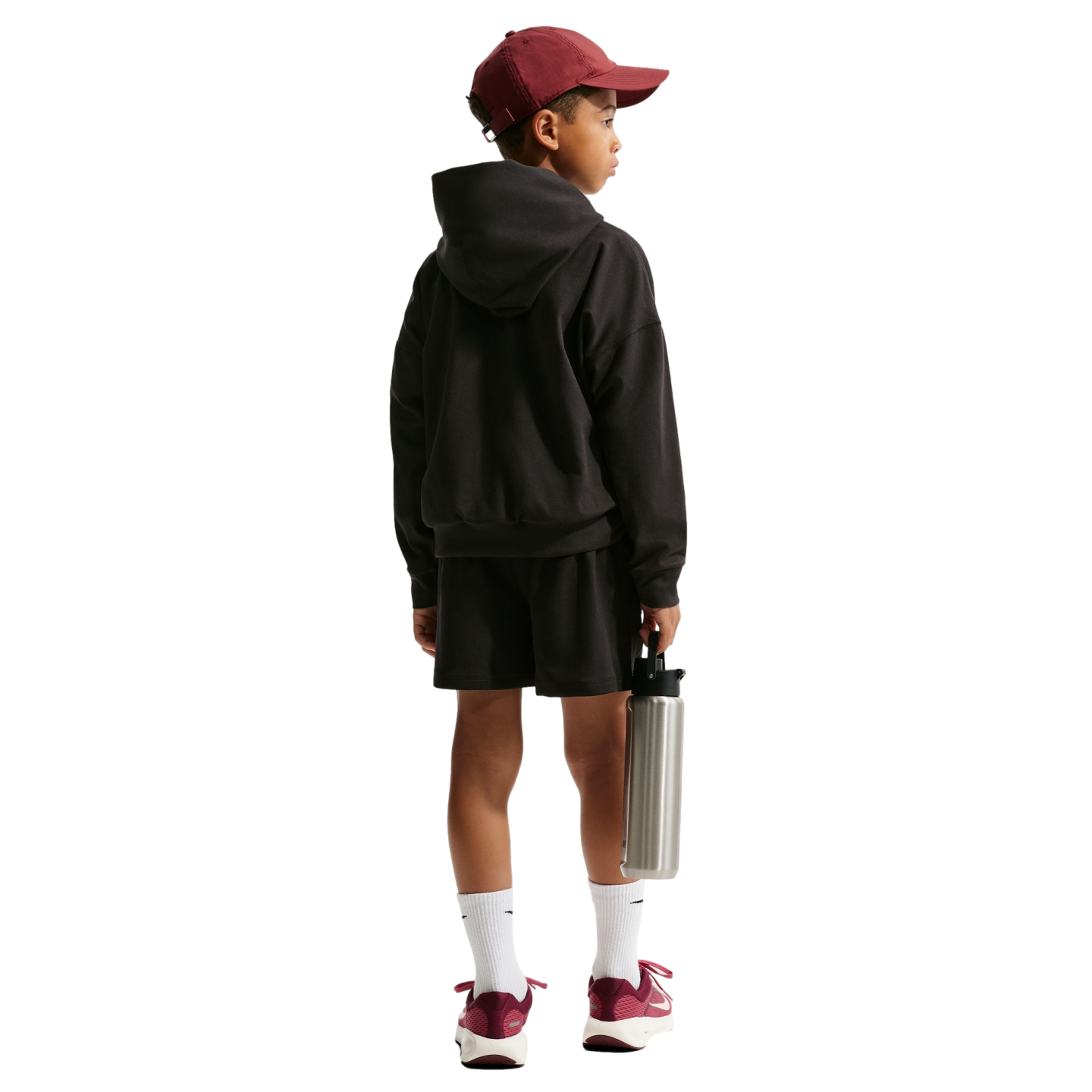 Back view of a child in a maroon cap, black hoodie, and shorts. They hold a silver water bottle and wear maroon sneakers against a black background.