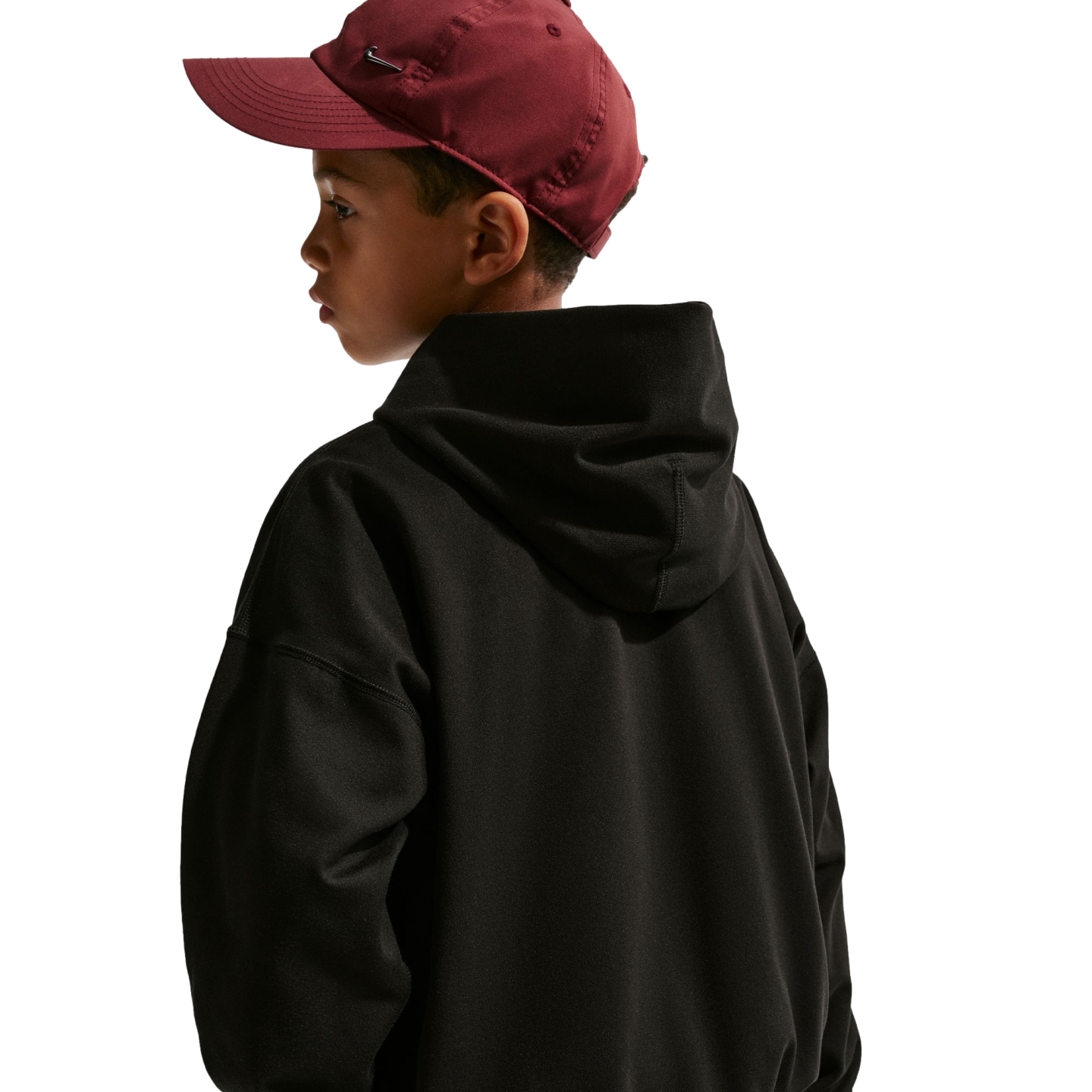 Side view of a young boy looking over his shoulder, wearing a maroon baseball cap and a black hoodie against a plain black background.