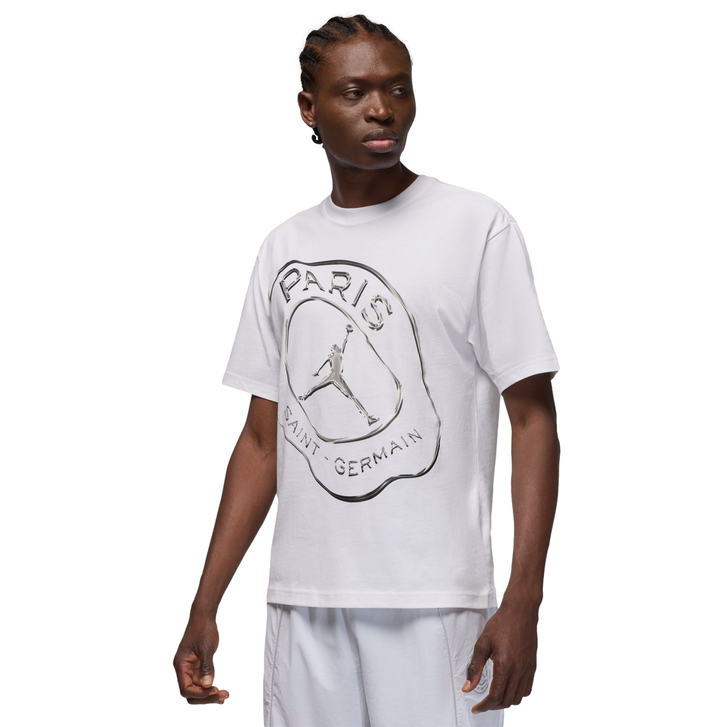 A person with braided hair wears a white T-shirt featuring a silver Jordan x PSG graphic, paired with white shorts against a solid black background.