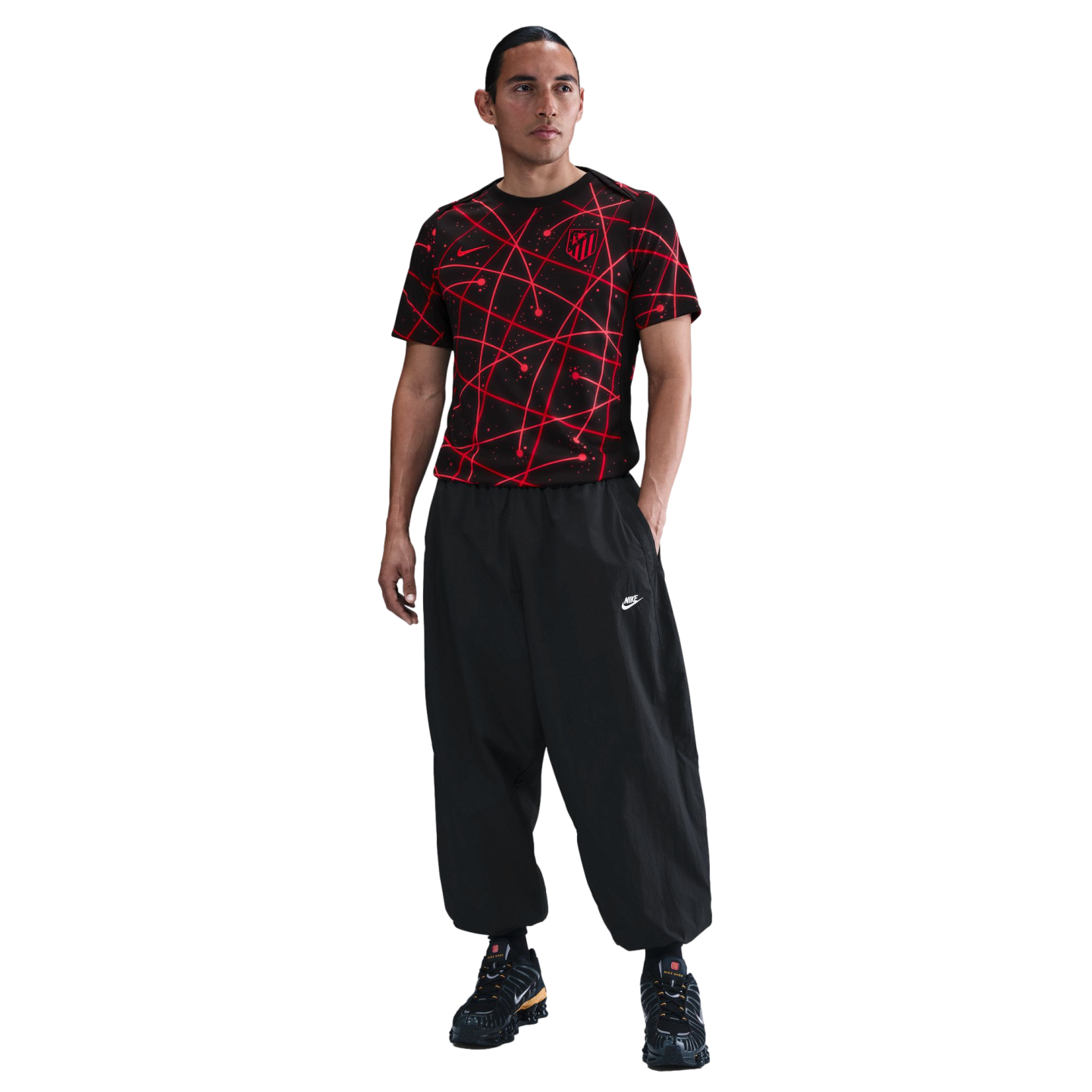 A man in a red and black patterned soccer jersey, baggy black pants, and sneakers, standing against a white background.