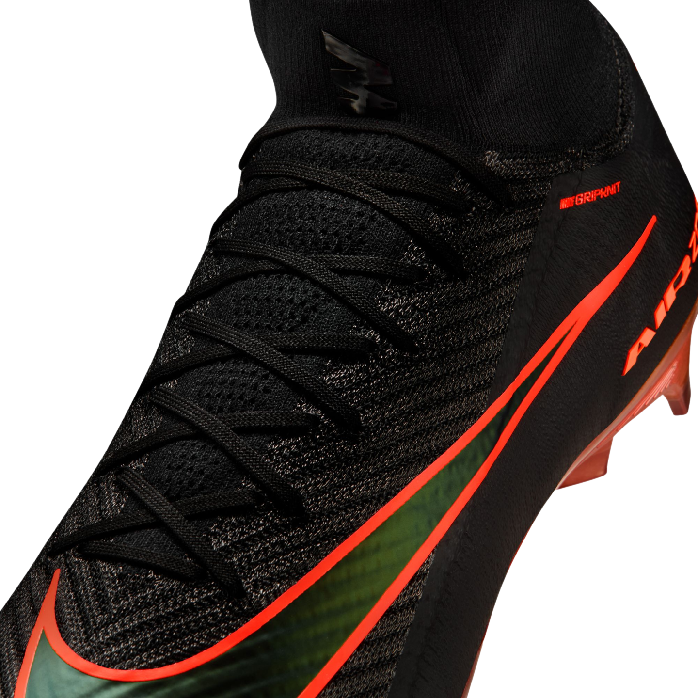 A close-up view of a black Nike soccer cleat with a textured knit upper and a sock-like collar. The shoe features a large, iridescent green Nike swoosh outlined in bright orange, along with small orange "NIKE GRIPKNIT" text and orange studs visible at the