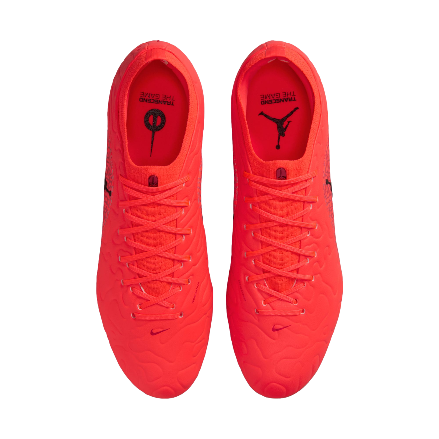 Top-down view of vibrant neon orange Nike and Jordan soccer cleats on a black background, with "TRANSCEND THE GAME" text on the insoles.