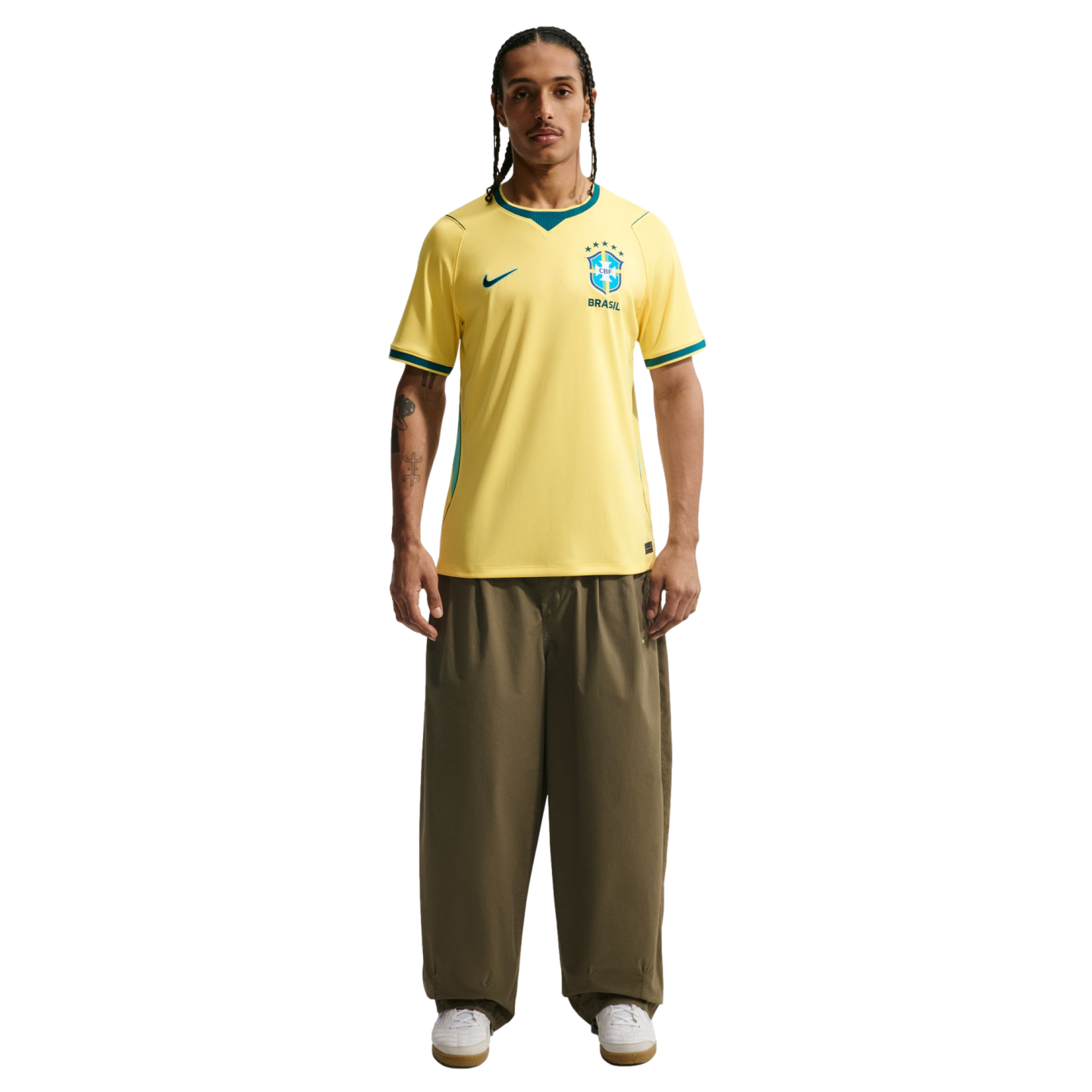 A man stands against a black background wearing a yellow Nike Brazil national team soccer jersey, olive green wide-leg trousers, and white sneakers. He has braided hair, a mustache, and tattoos on his right arm.