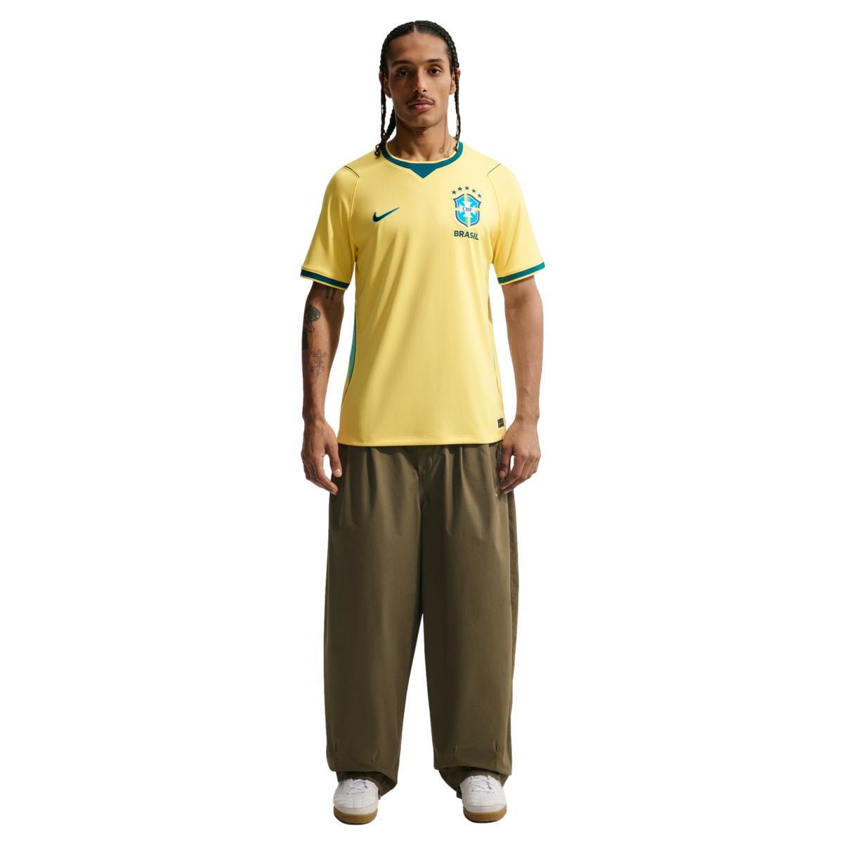 A man stands against a black background wearing a yellow Nike Brazil national team soccer jersey, olive green wide-leg trousers, and white sneakers. He has braided hair, a mustache, and tattoos on his right arm.